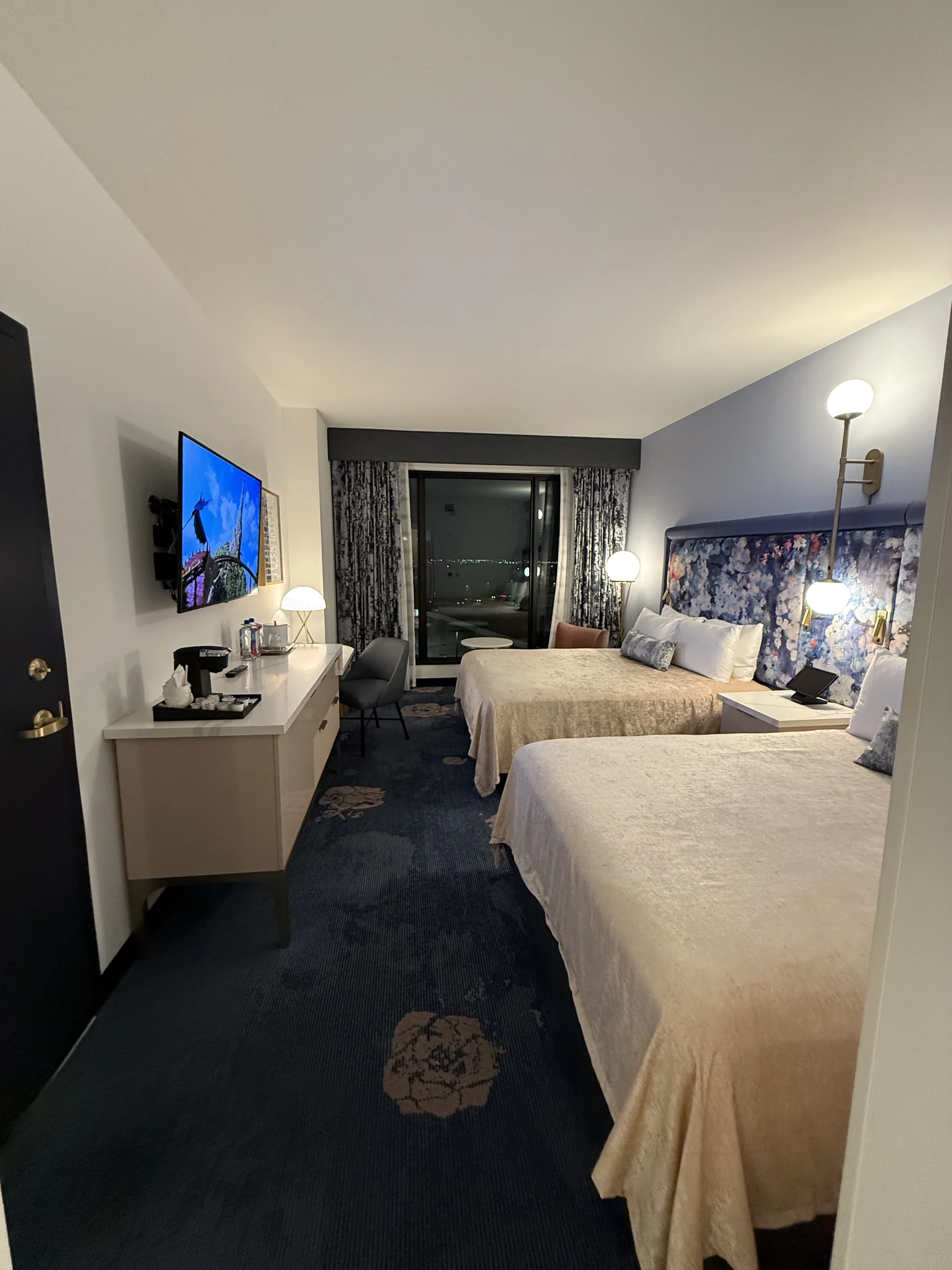 A hotel room with two queen beds, floral headboards, bedside lamps, a wall-mounted television, a desk, and a balcony with city lights visible outside.