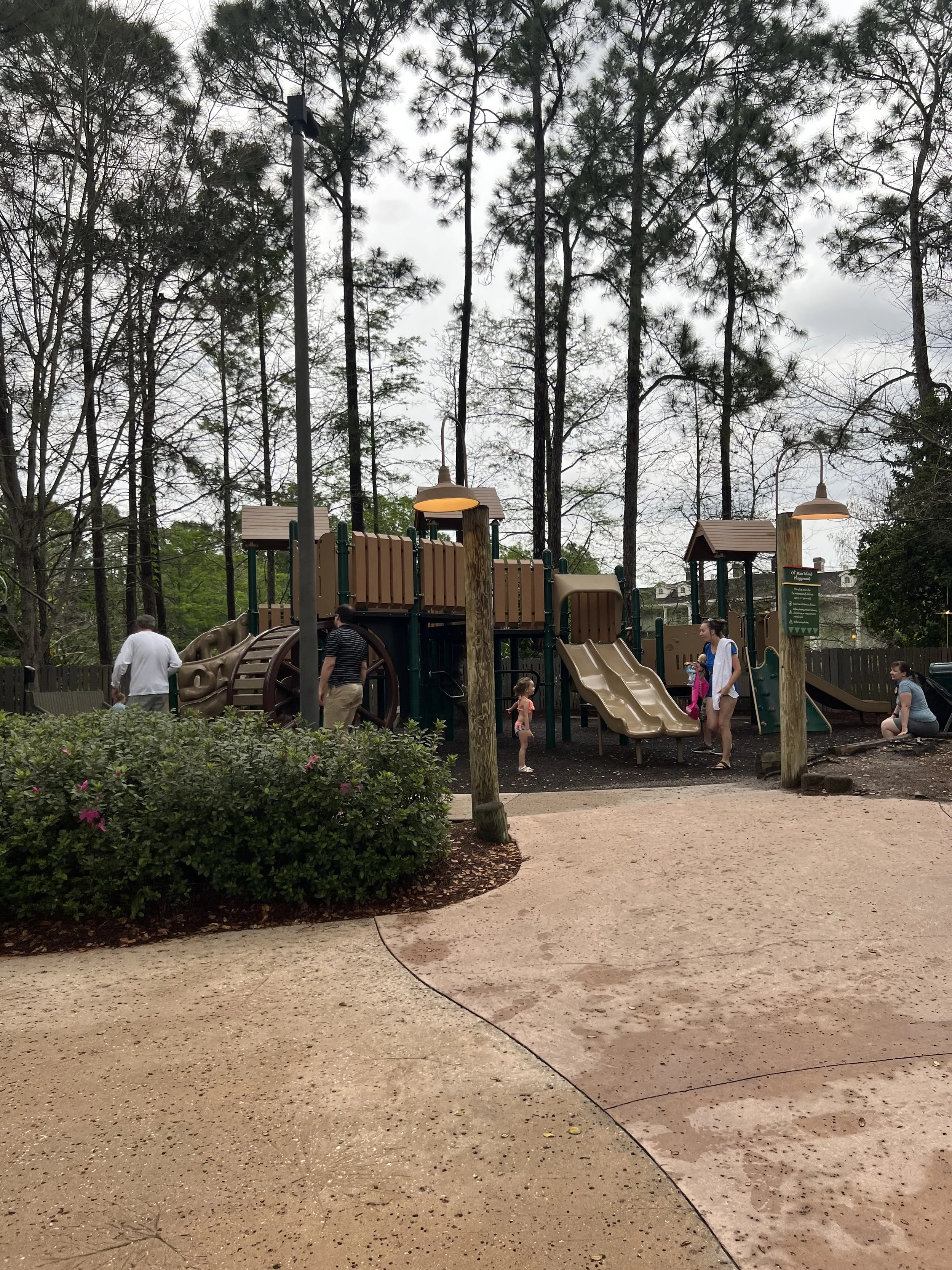 Children and adults playing on a jungle gym with slides at a playground surrounded by trees.