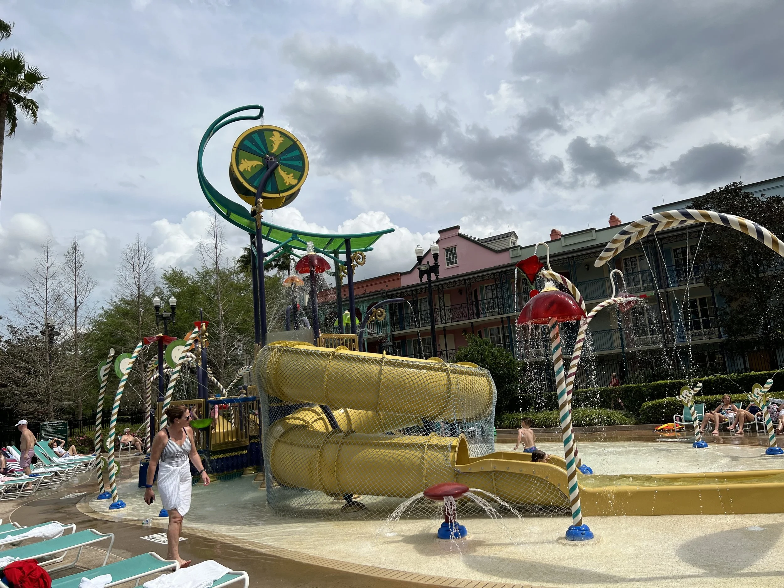 People enjoying a water play area with slides, water sprays, and colorful umbrellas, surrounded by lounge chairs, trees, and a pastel-colored building under a cloudy sky.