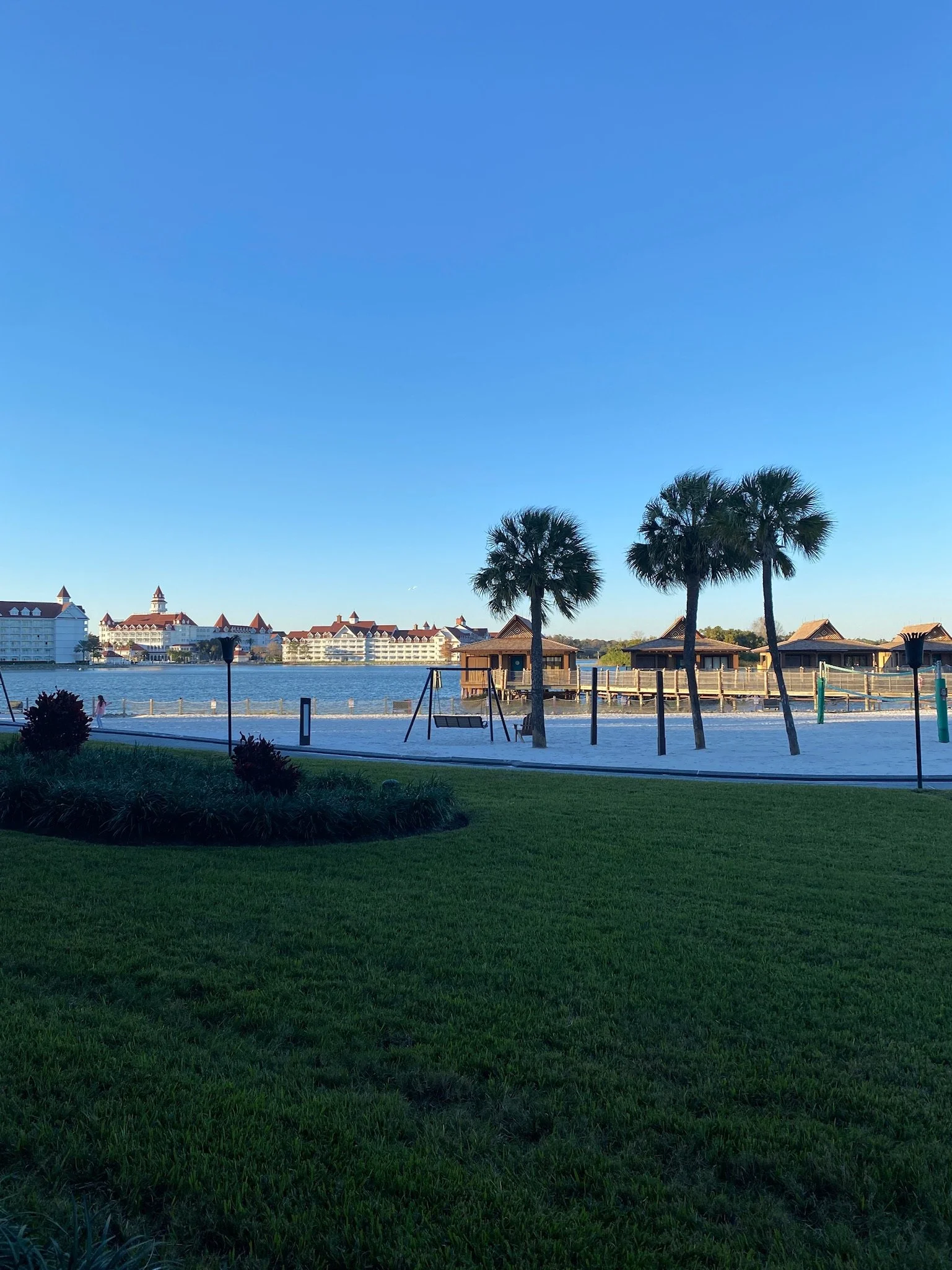 View of a grassy area and palm trees near a waterfront with bungalows, a swing set, and a large hotel or resort building in the background under a clear blue sky.