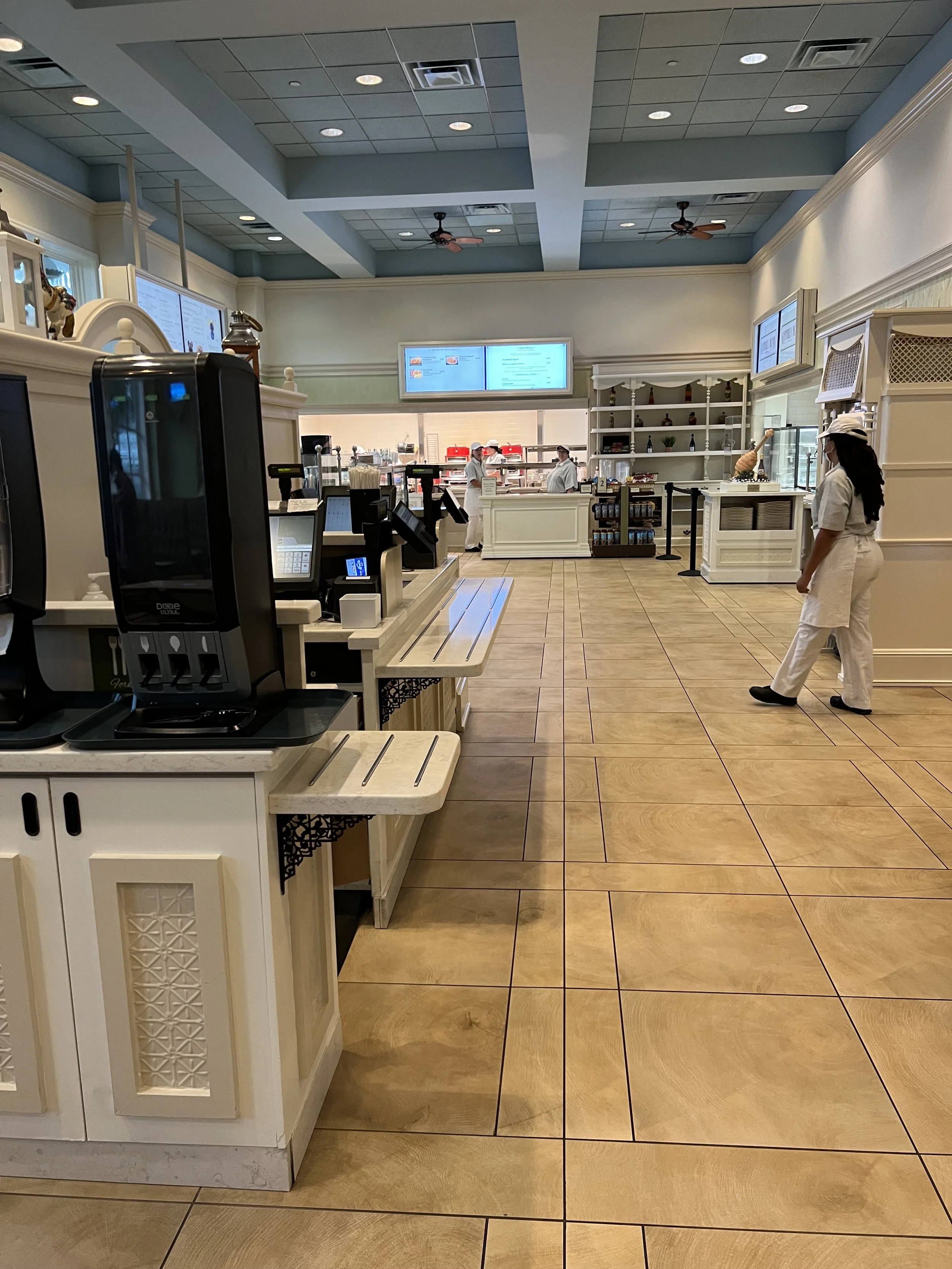 Interior of a restaurant or café with a counter area in the foreground, staff members in white uniforms working behind the counter, digital menu screens on the wall, and a customer dressed in white and wearing a face mask waiting or walking by.