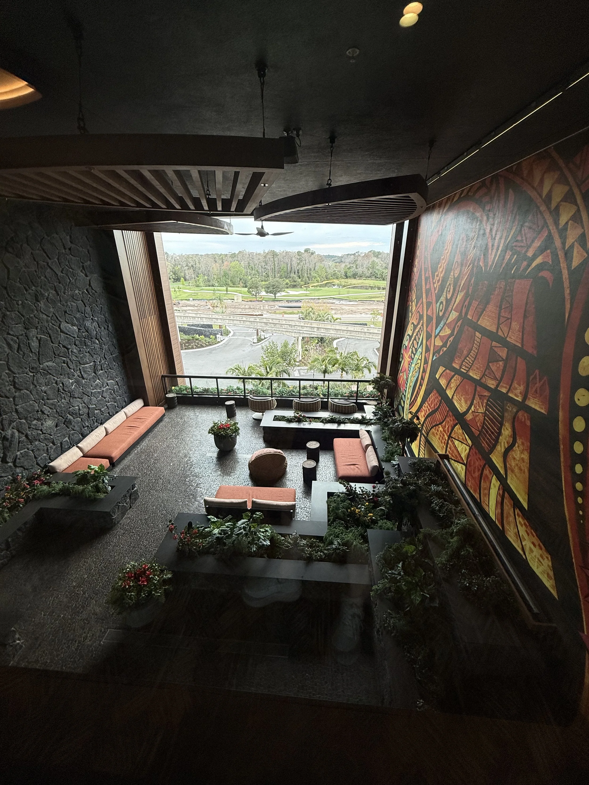 Indoor lounge area with orange cushions on benches and chairs, a large window overlooking a golf course, and decorative plants and flowers.