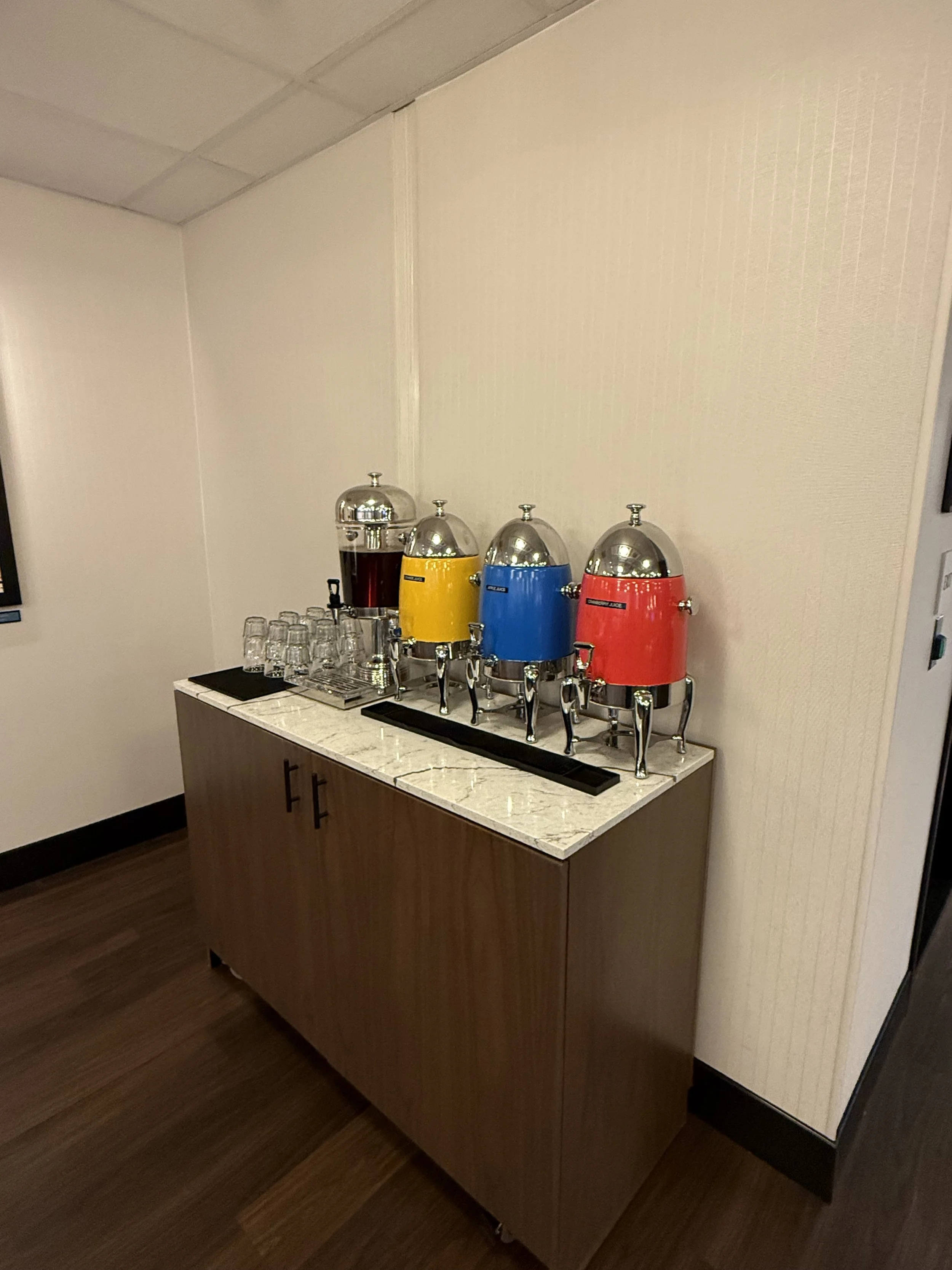 A juice and beverage station with four large dispensers in red, blue, yellow, and black, placed on a white marble counter. Several glasses are stacked beside the dispensers.
