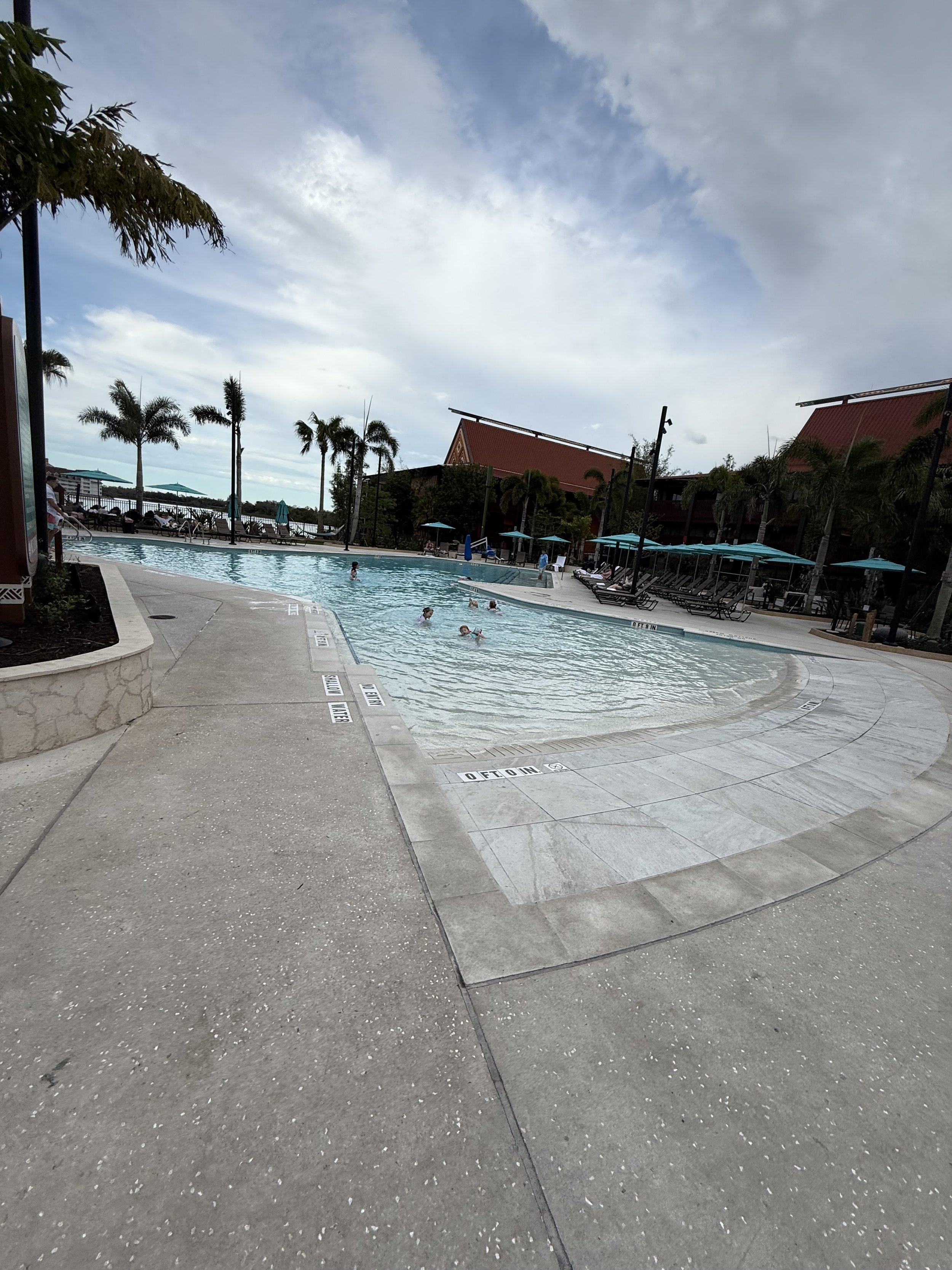A swimming pool area with a few people swimming, surrounded by palm trees, lounge chairs, and shaded cabanas under a partly cloudy sky.
