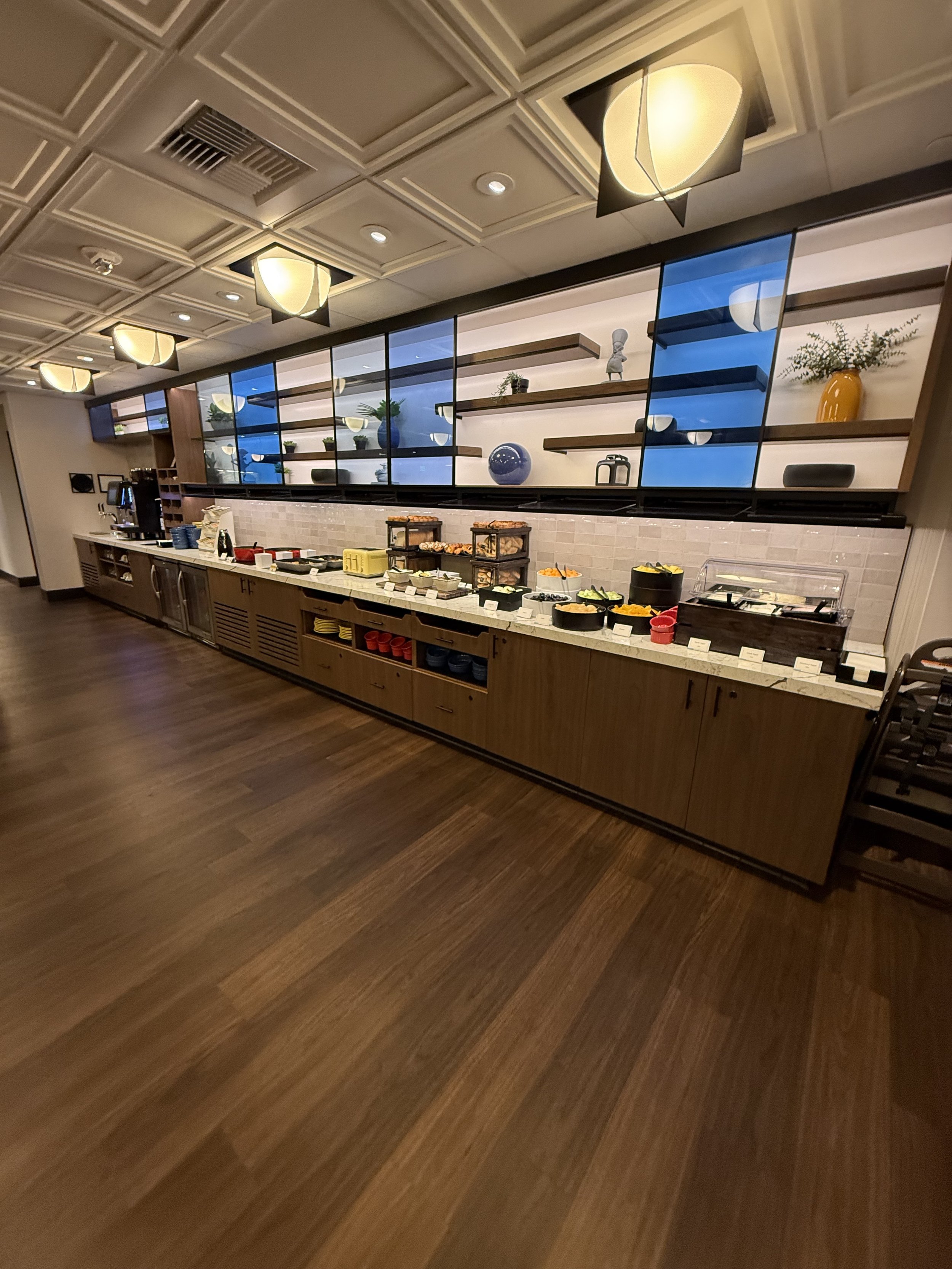 Buffet breakfast area with various food options, bowls, and condiment containers on a long counter, behind which are open shelves with decorative items, such as vases and plants, and a large mirror reflecting ceiling lights.