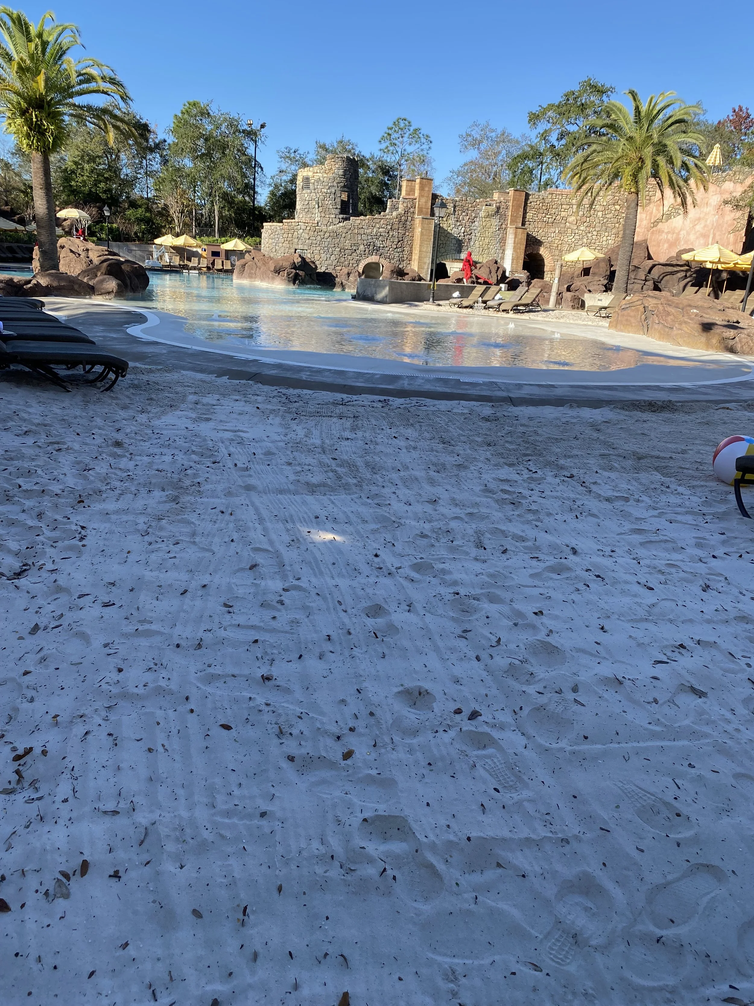 Empty swimming pool area with lounge chairs, umbrellas, palm trees, and rocky structures against a blue sky.