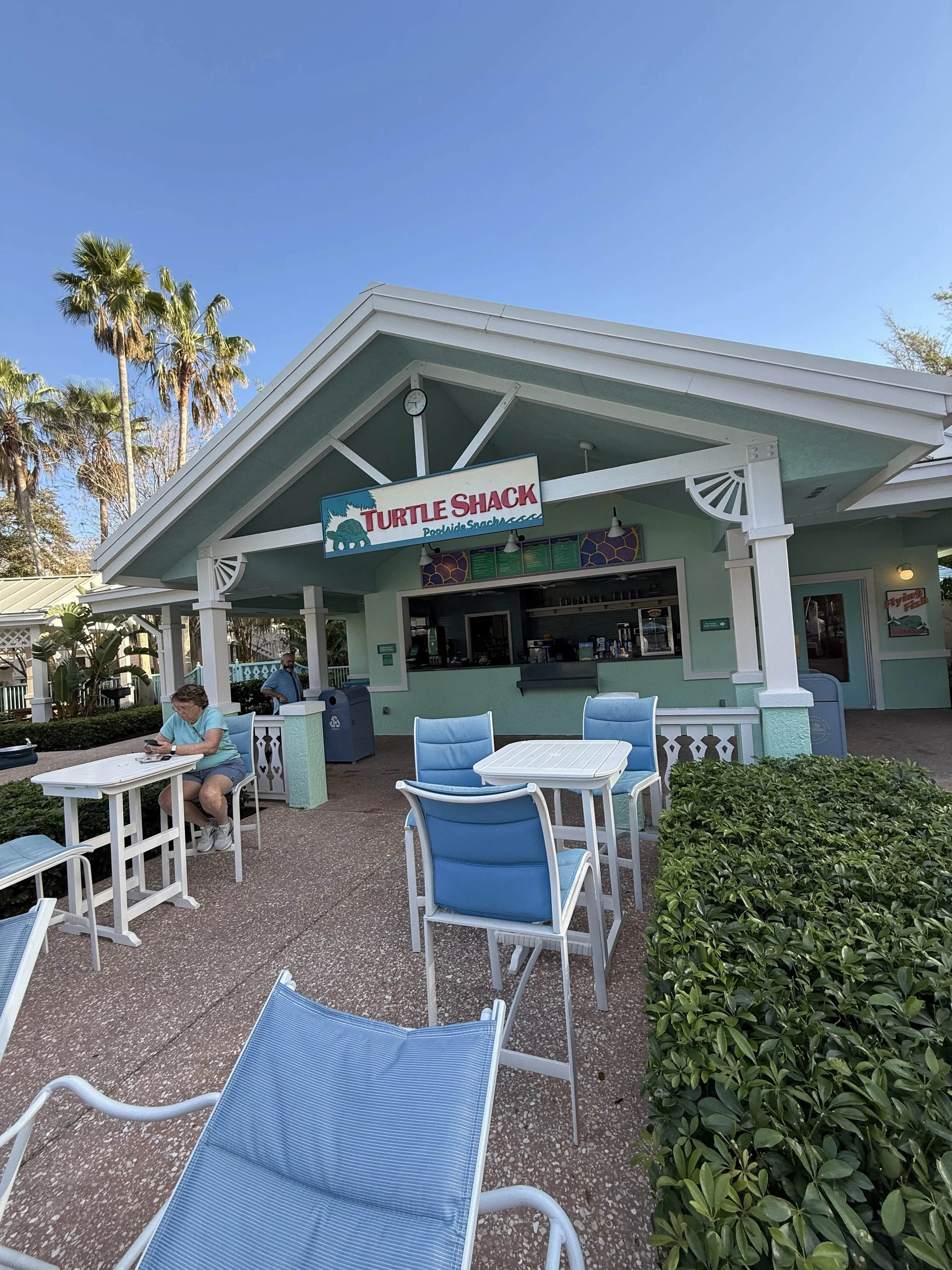 The exterior of a Turtle Shack restaurant with outdoor seating, green building, blue chairs, and palm trees in the background.