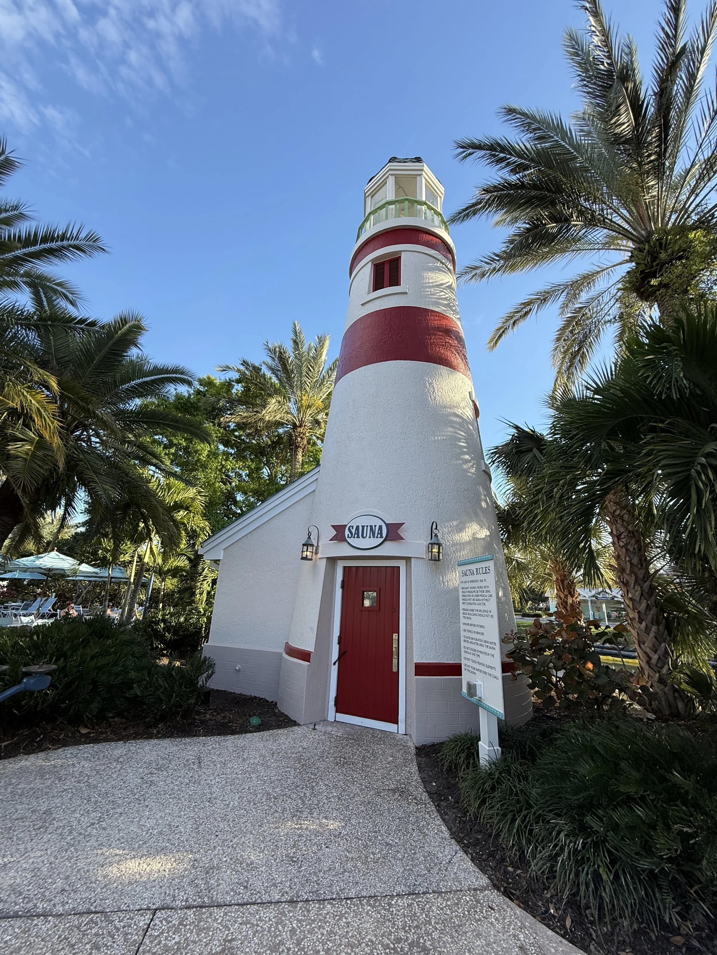 A tall, white lighthouse with red horizontal stripes, surrounded by palm trees and greenery, with a clear blue sky in the background. The lighthouse has a small building at its base labeled 'Sauna' with a red door and two lantern-style lights. There 