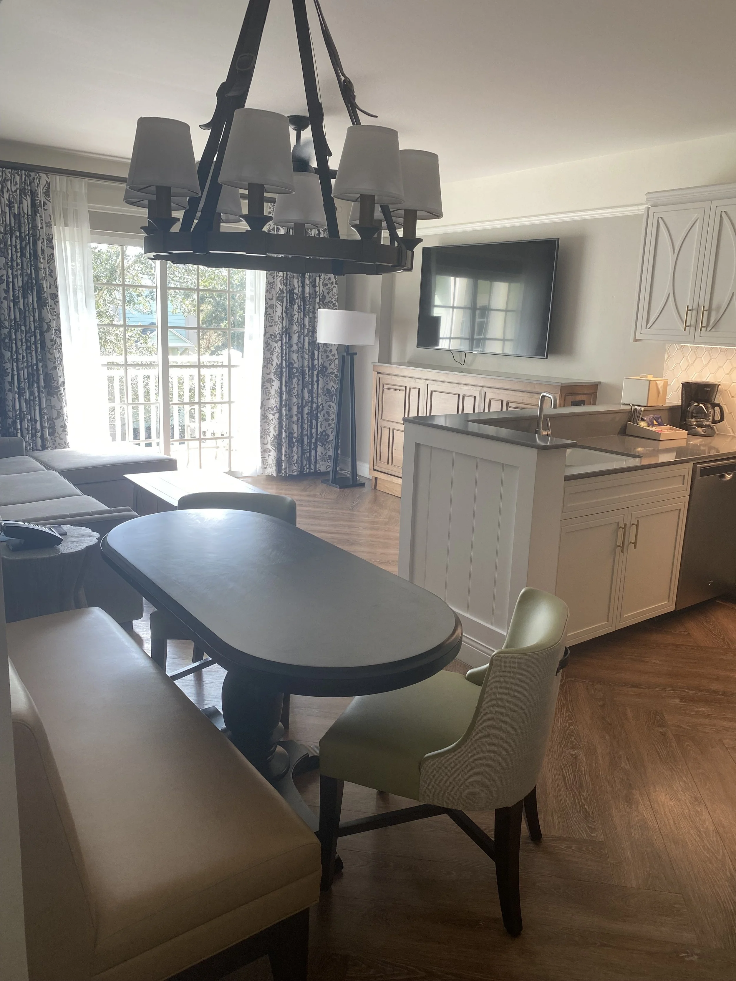 Living room with sliding glass door, curtains, TV, wooden cabinet, lamp, kitchen with sink and coffee maker, table with chairs.