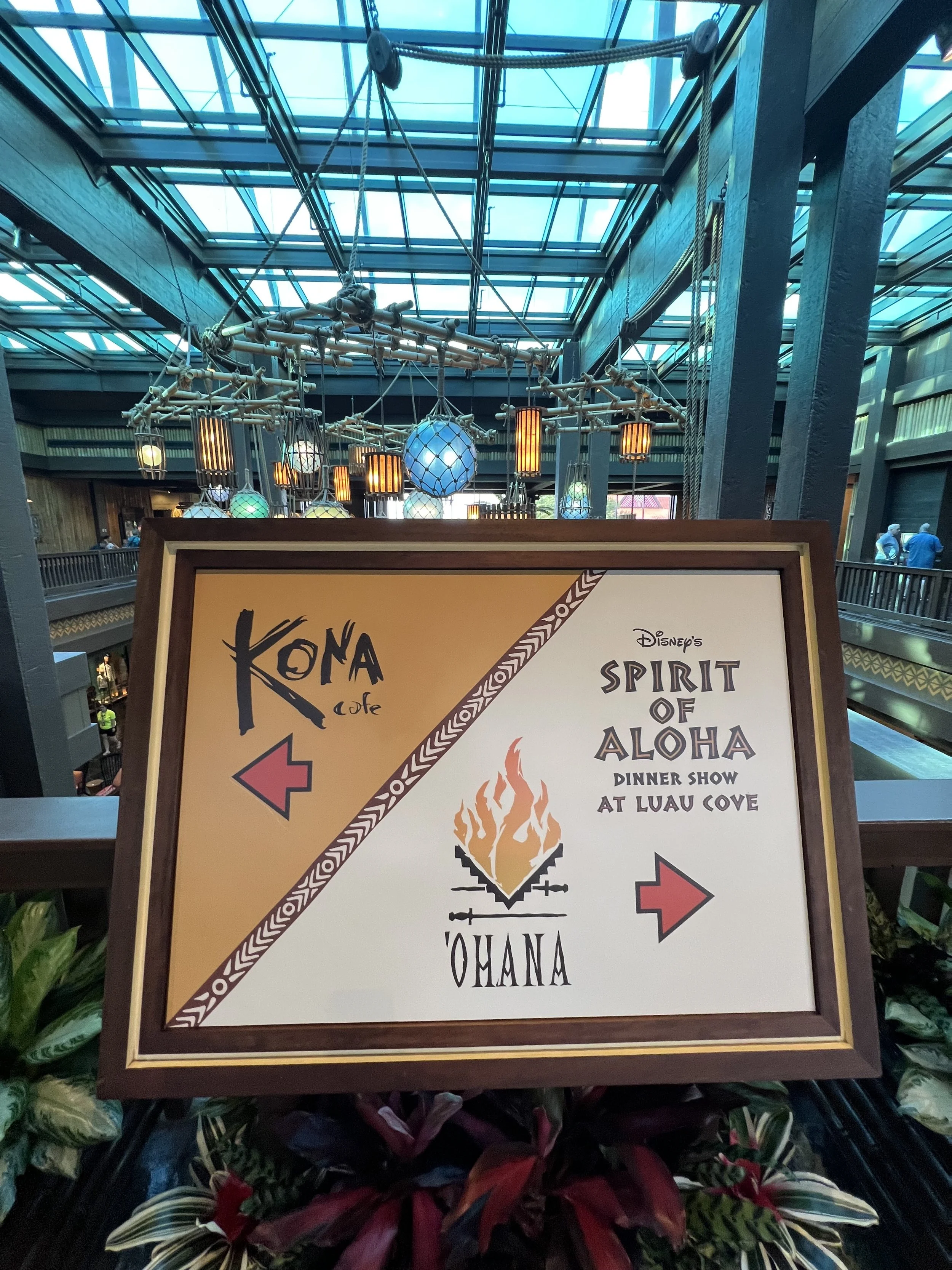 Indoor shopping area with a sign directing to Kona Cafe on the left and O'Hana with a fire graphic on the right, under a glass ceiling with hanging lights and decorative elements.