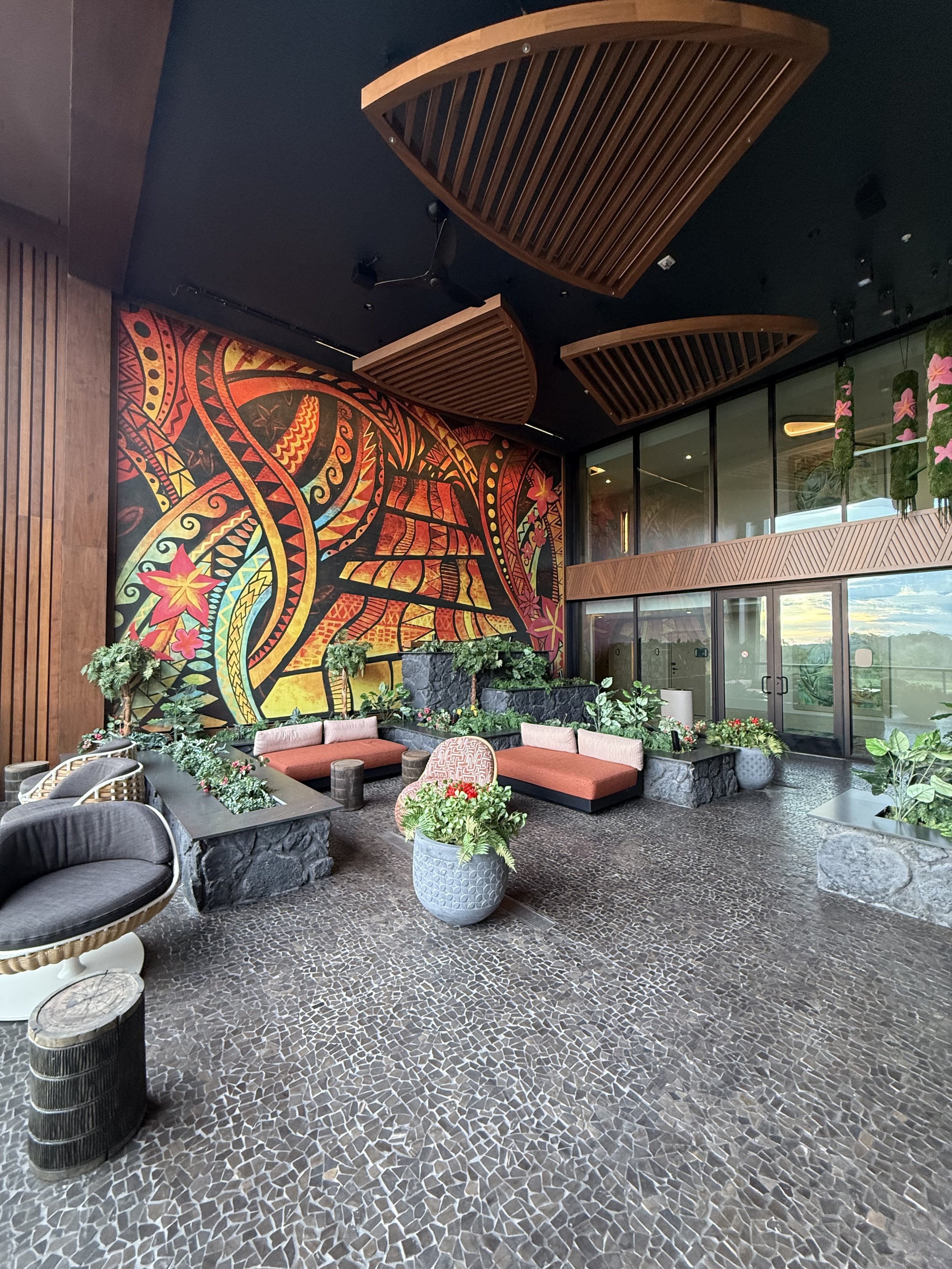 Indoor lounge area with mosaic floor, plants, and colorful wall mural with tropical and tribal patterns. There are built-in seating with cushions and decorative wood ceiling panels.