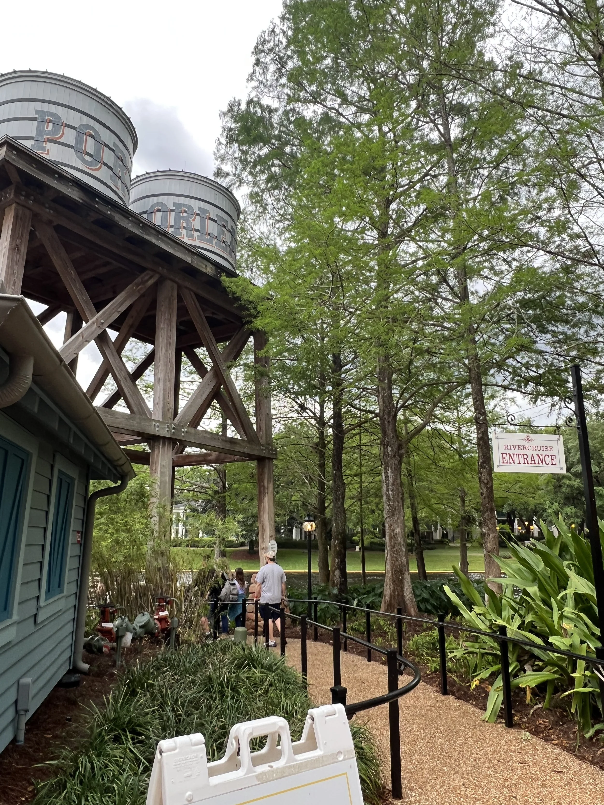A pathway near trees leads to an entrance sign for Rivercruise. A man and children walk along the path near water towers labeled "POPE" in the background.