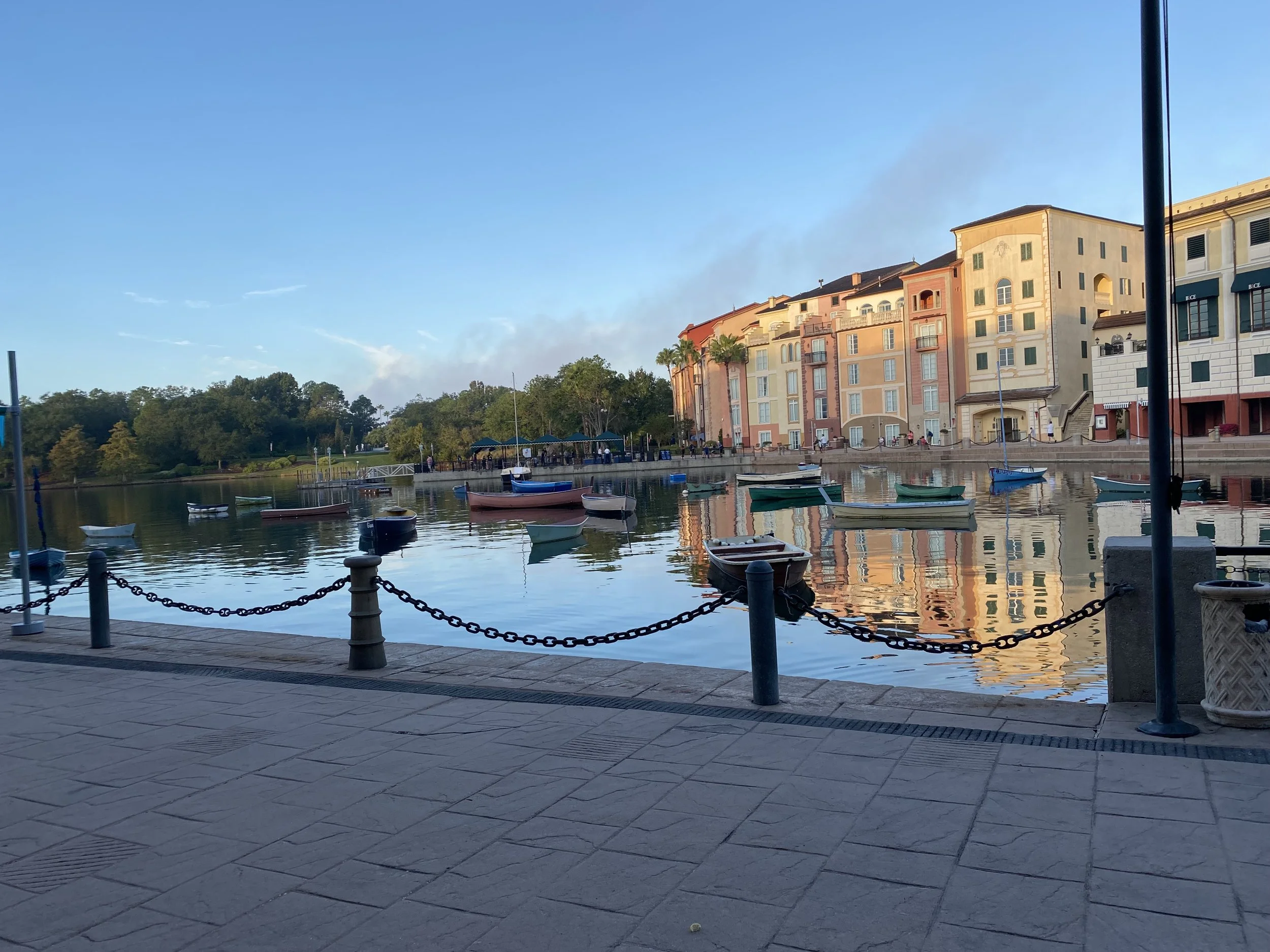 A tranquil waterway with several small boats anchored along the edge, adjacent to a walkway with chains and posts, and colorful multi-story buildings on the opposite bank reflecting in the water, under a clear blue sky.
