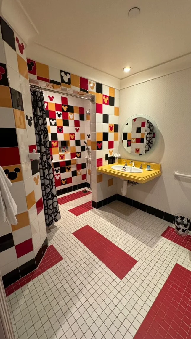 A colorful bathroom with a shower area on the left, featuring black, red, white, and yellow Mickey Mouse-themed tiles and a black and white patterned shower curtain. The right side has a yellow sink countertop with a round mirror above, a small flowe
