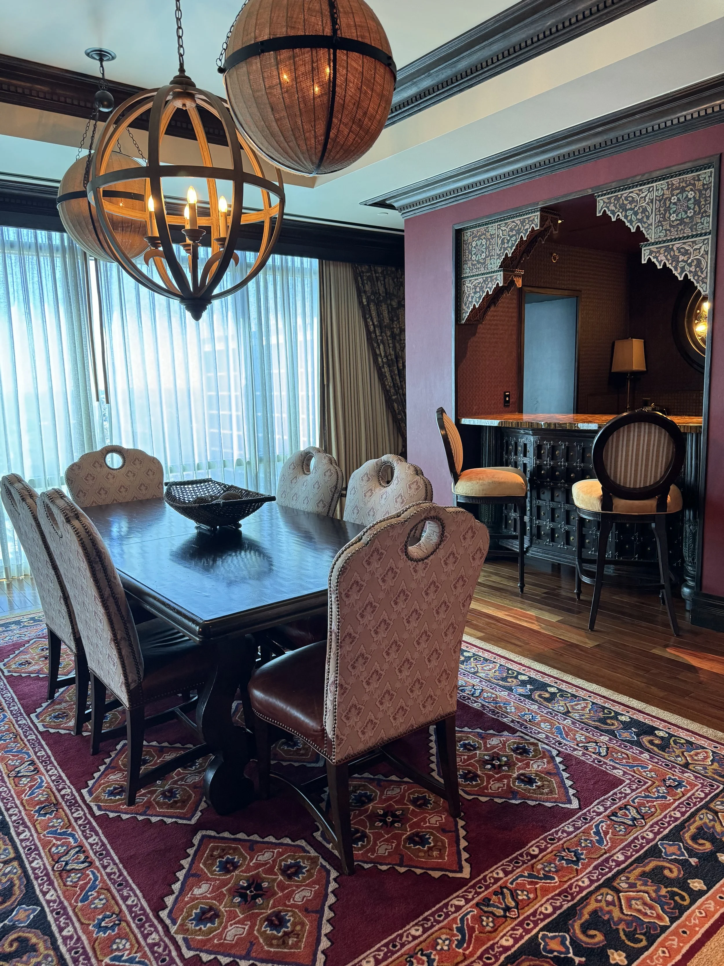 Elegant dining room with a large wooden table, beige patterned chairs, a colorful Persian rug, large windows with sheer blue curtains, a bar area with two high chairs, and a chandelier with spherical wood design