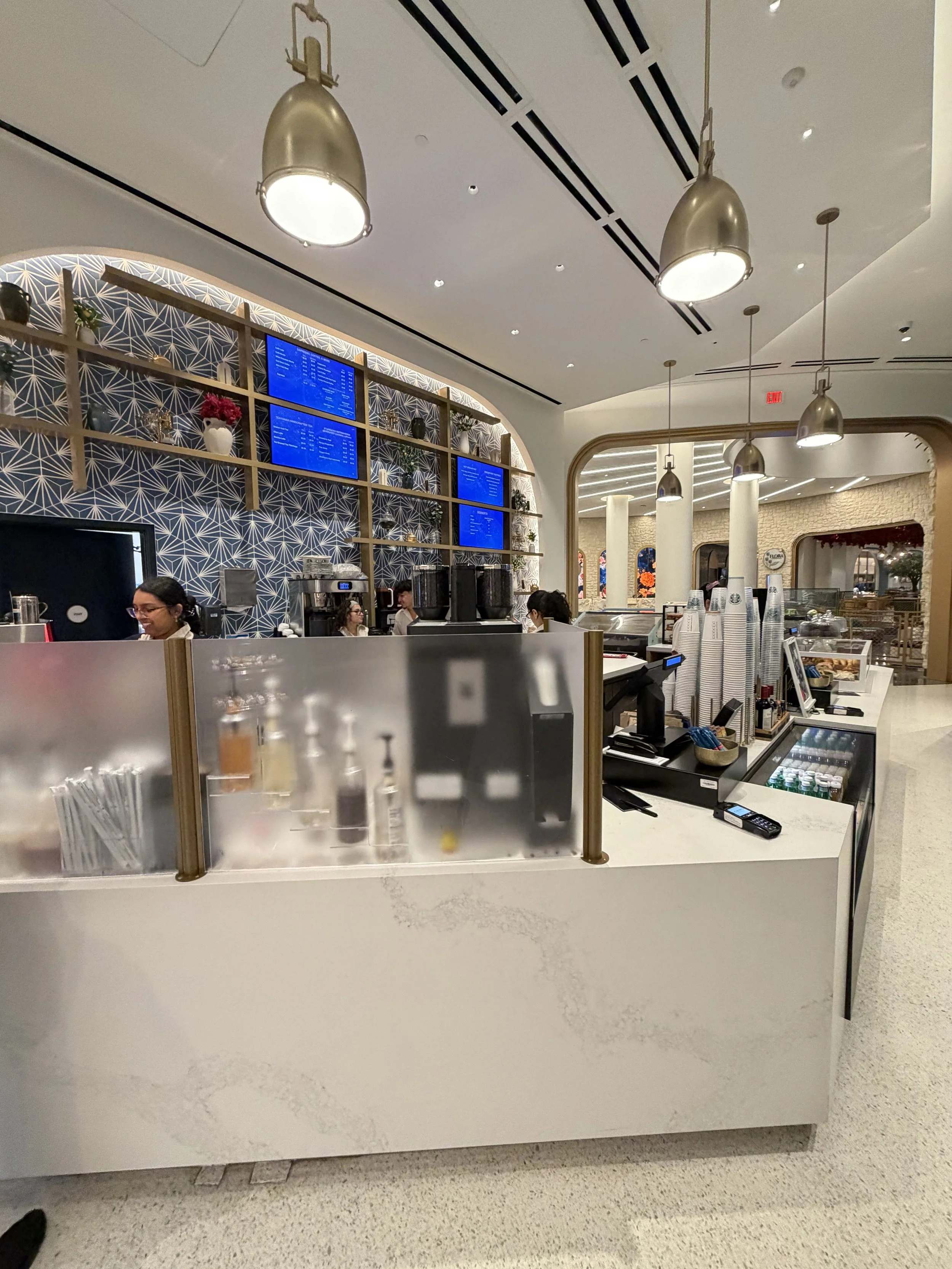 A modern cafe counter with coffee and beverage equipment, stacked paper cups, and digital screens displaying the menu, inside a stylish, well-lit space.