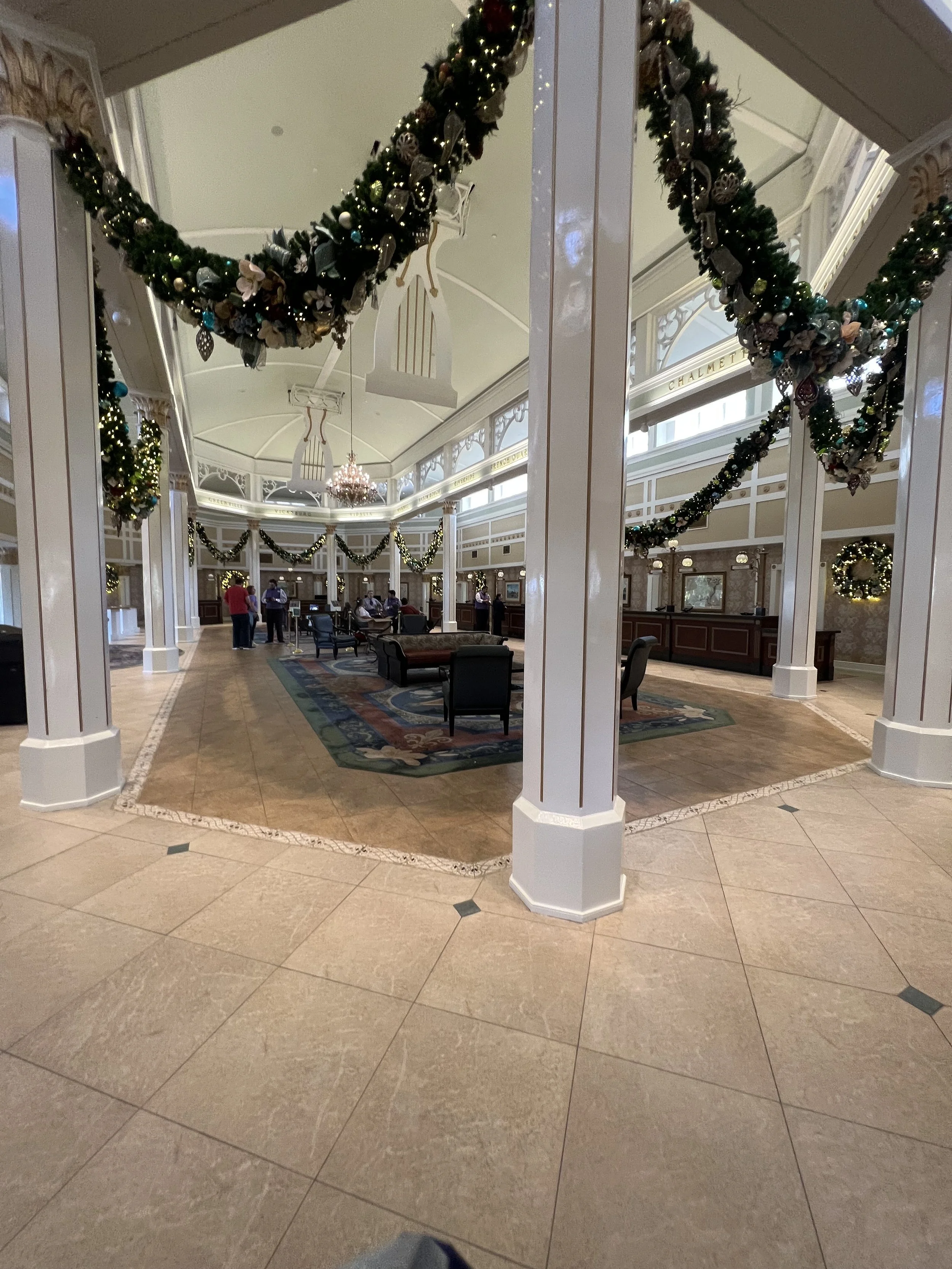 The interior of a grand hotel lobby decorated for Christmas with garlands, ornaments, and lights hanging from the ceiling and around the pillars. There are people in the background near a check-in desk and seating areas.