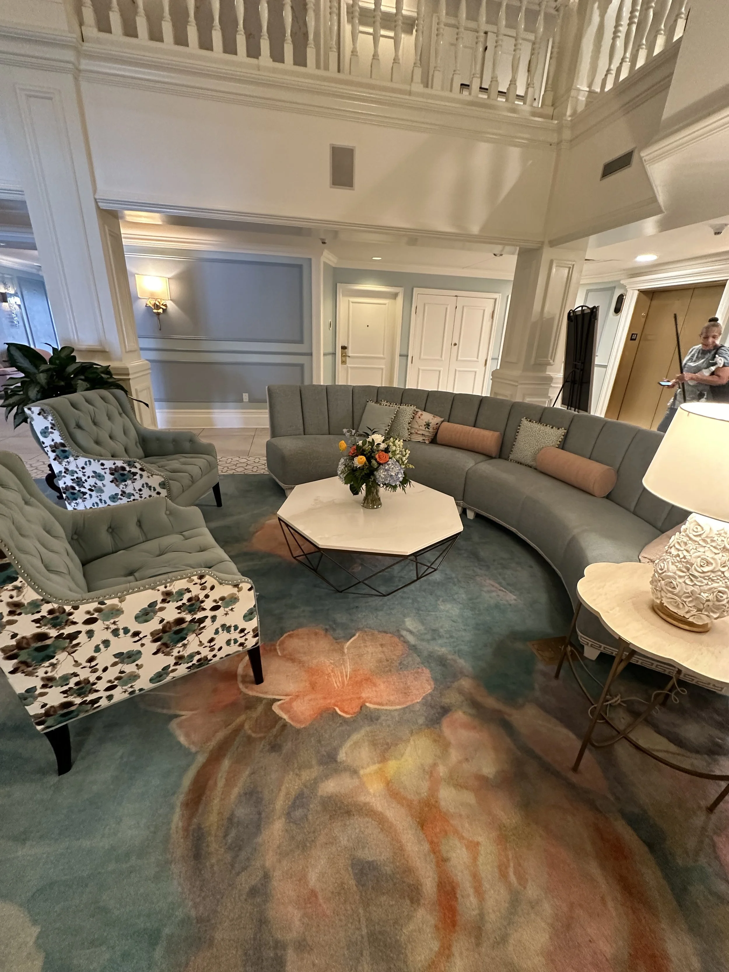Elegant living room with curved gray sofa, floral armchairs, colorful floral rug, and a white octagonal coffee table with a flower arrangement, in a well-lit space with white paneling and a balcony.