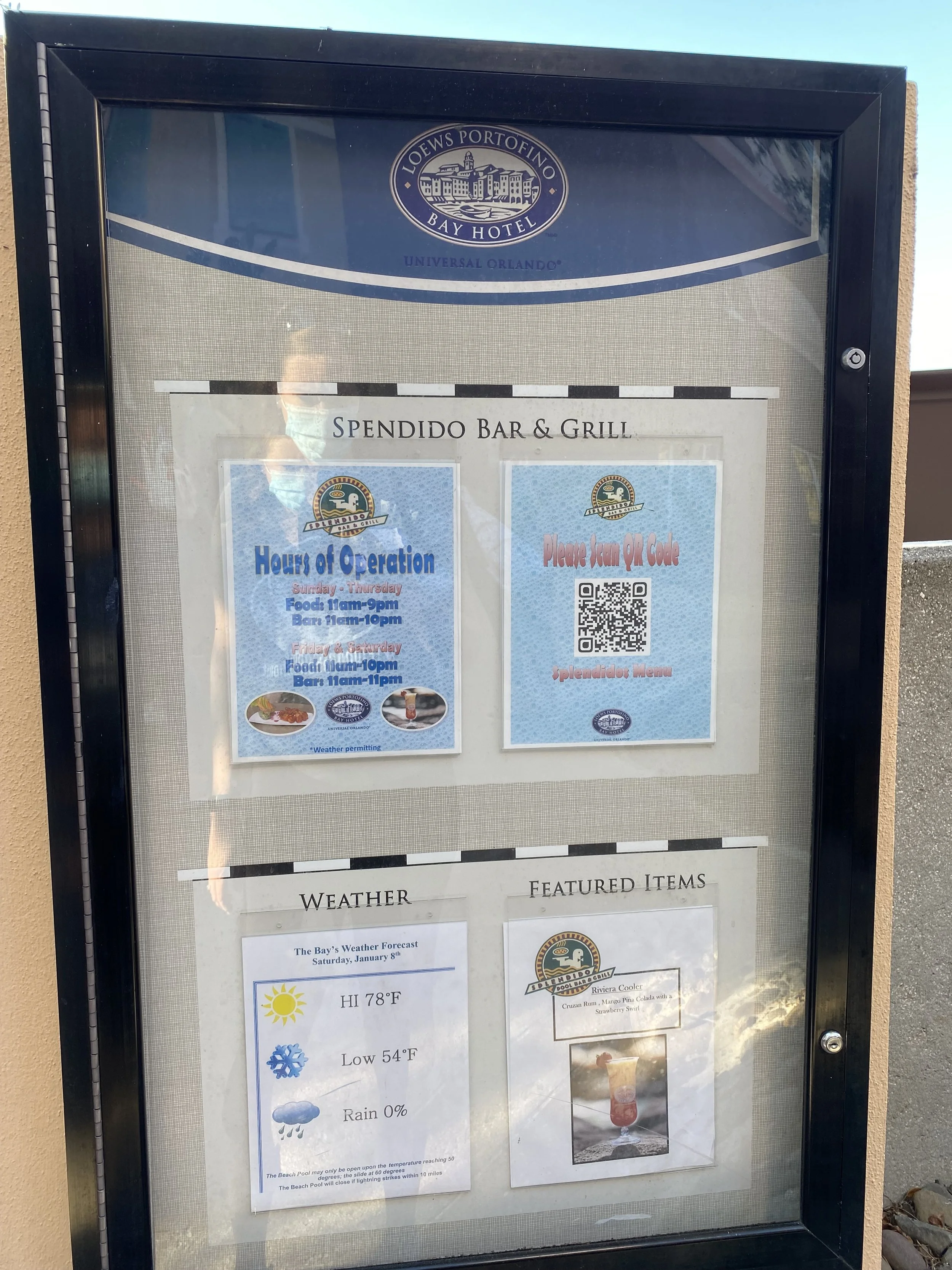 A restaurant menu display board outside at the Lowes Portofino Bay Hotel in Universal Orlando. The board shows the hours of operation for Spendido Bar & Grill, weather forecast details, and featured menu items, with a QR code for the menu.