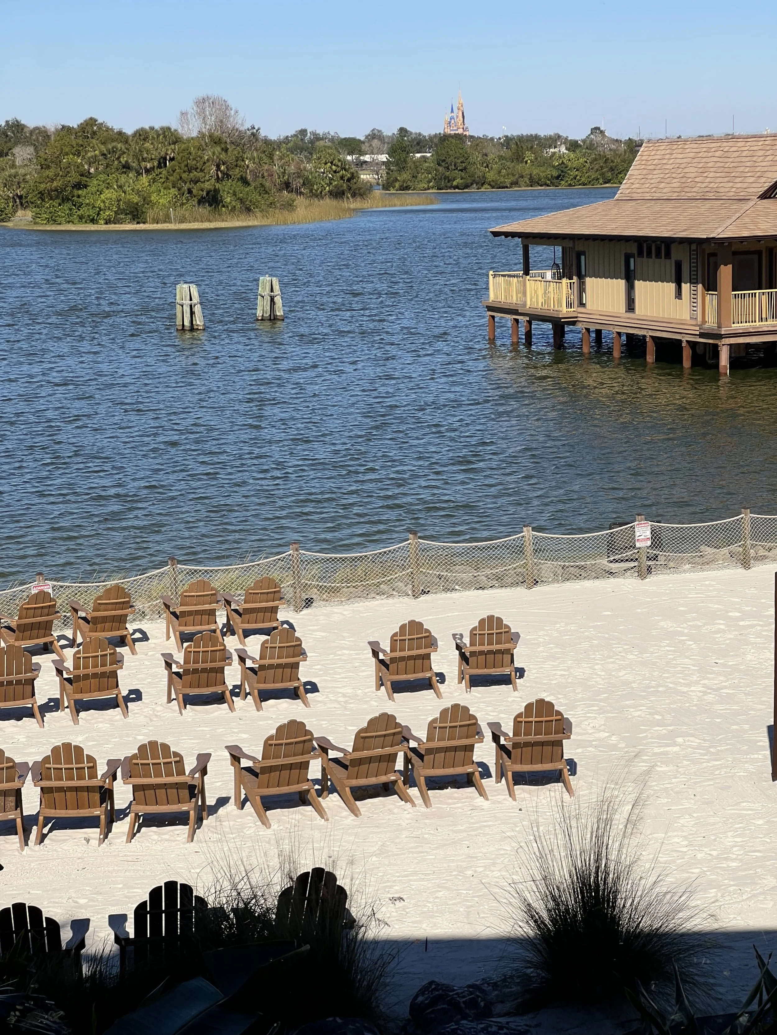 A beach scene with multiple wooden chairs arranged in rows on white sand, overlooking a body of water, with a wooden pier and house on stilts in the background, and trees along the shoreline.