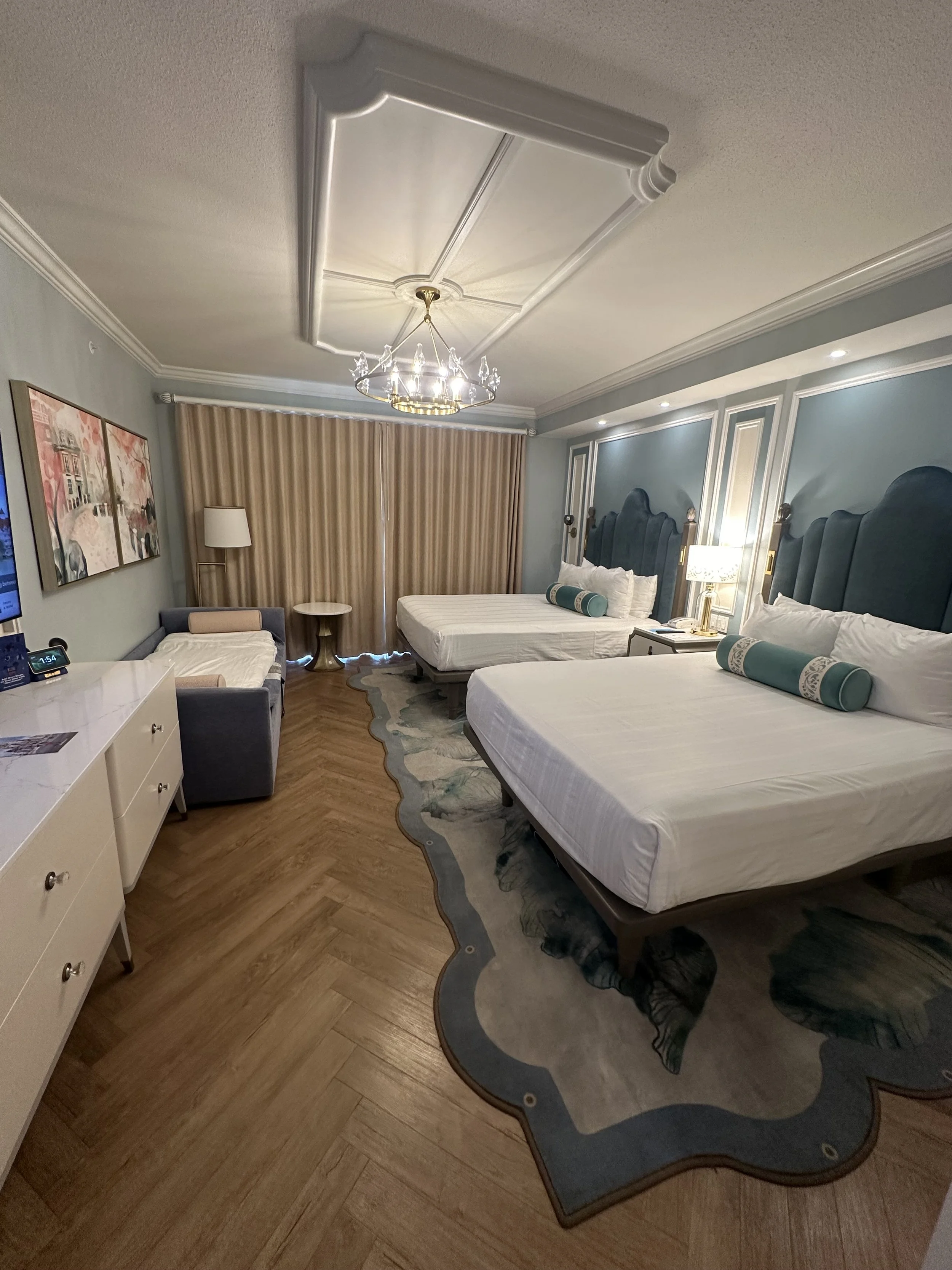 A hotel room with two beds, a chandelier, a TV, a small sofa, nightstand, and a window with beige curtains.