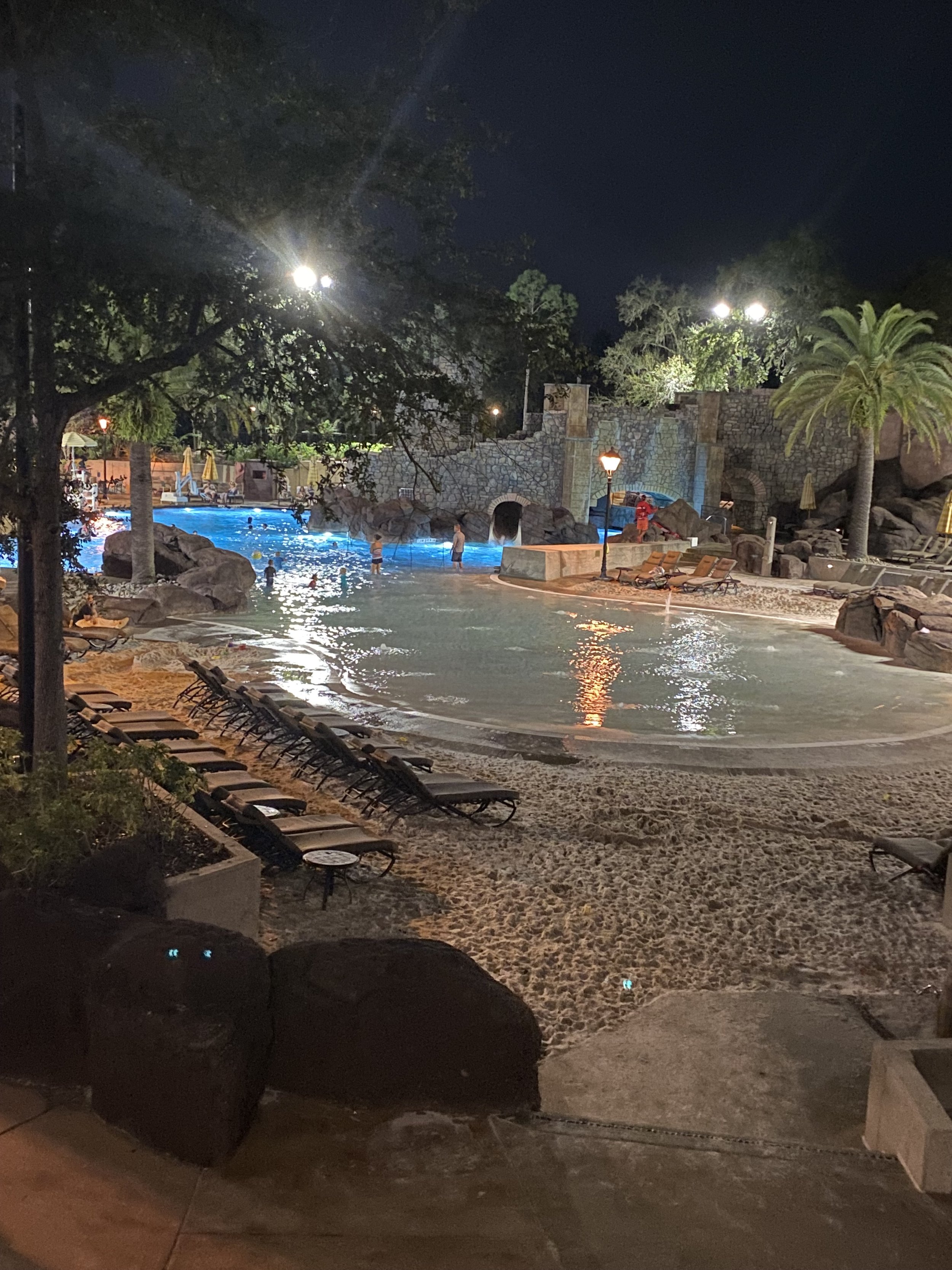 Nighttime scene of a swimming pool with people in the water, surrounded by lounge chairs, rocks, and palm trees, illuminated by pool and lamp lights.