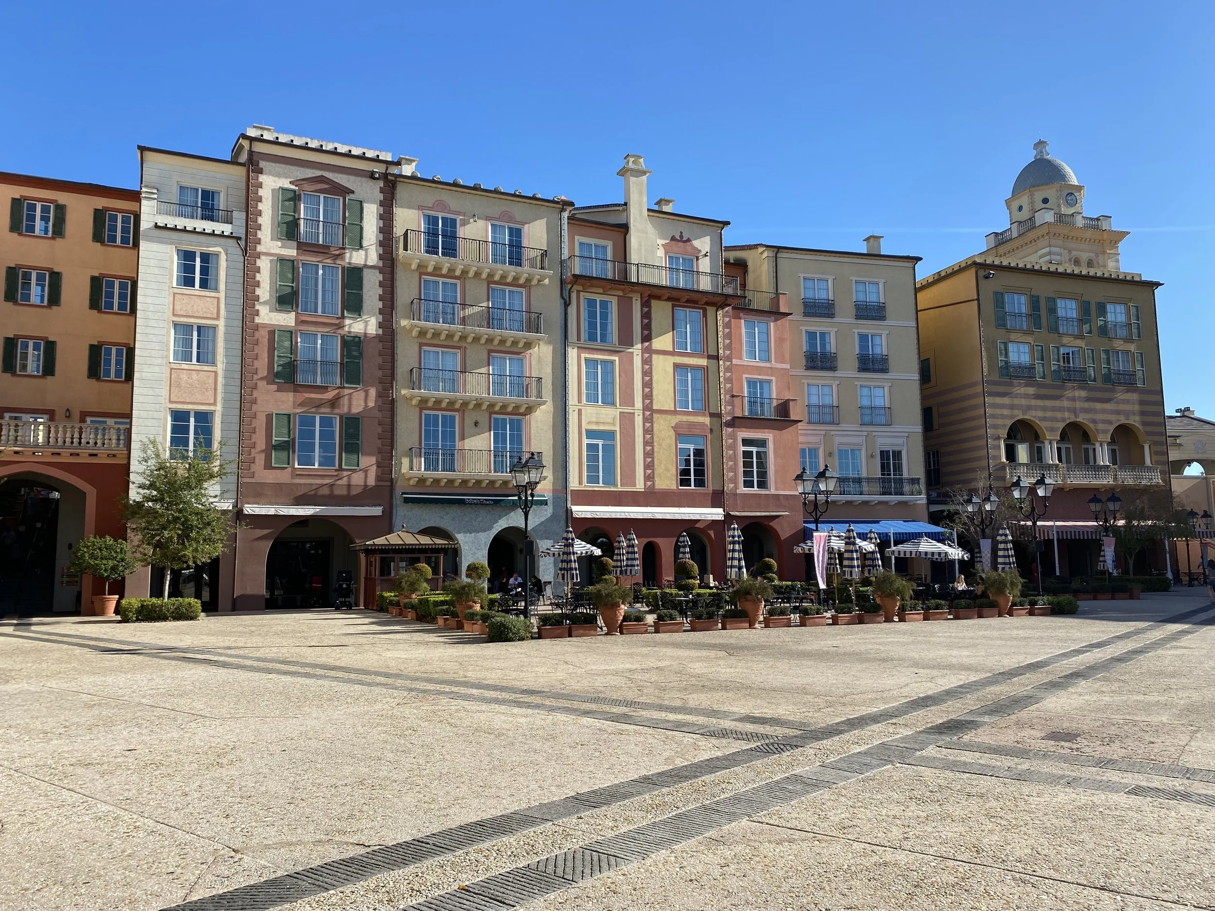 Colorful European-style buildings with balconies, storefronts, outdoor seating, and striped umbrellas, under a clear blue sky.