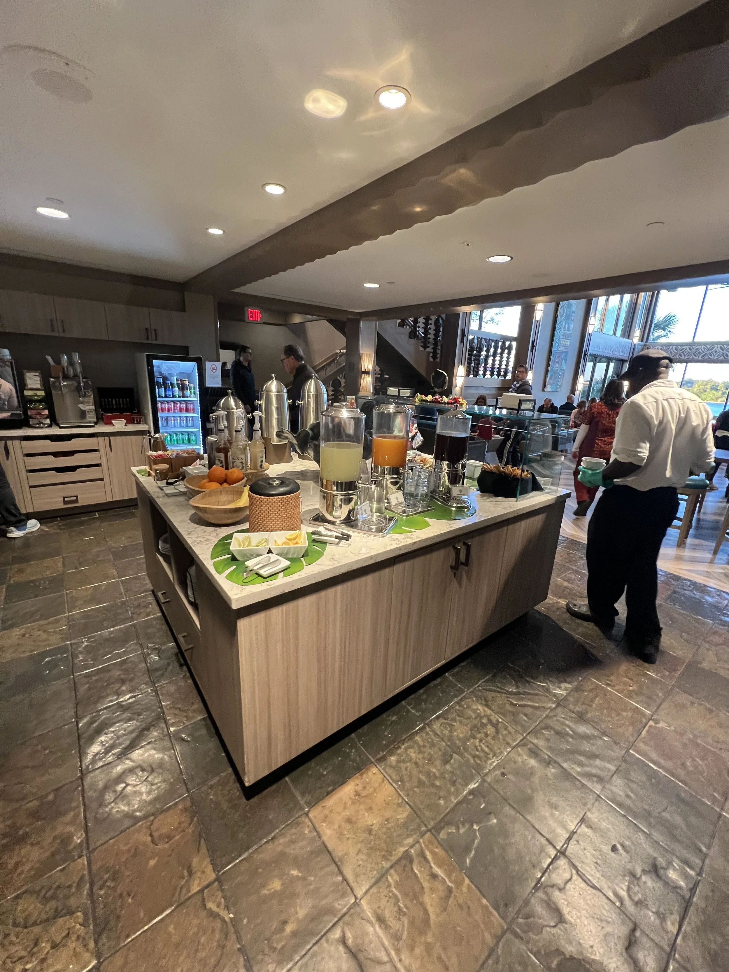 Buffet breakfast station with beverages, fruit, and breakfast items in a restaurant with large windows and several people in the background.