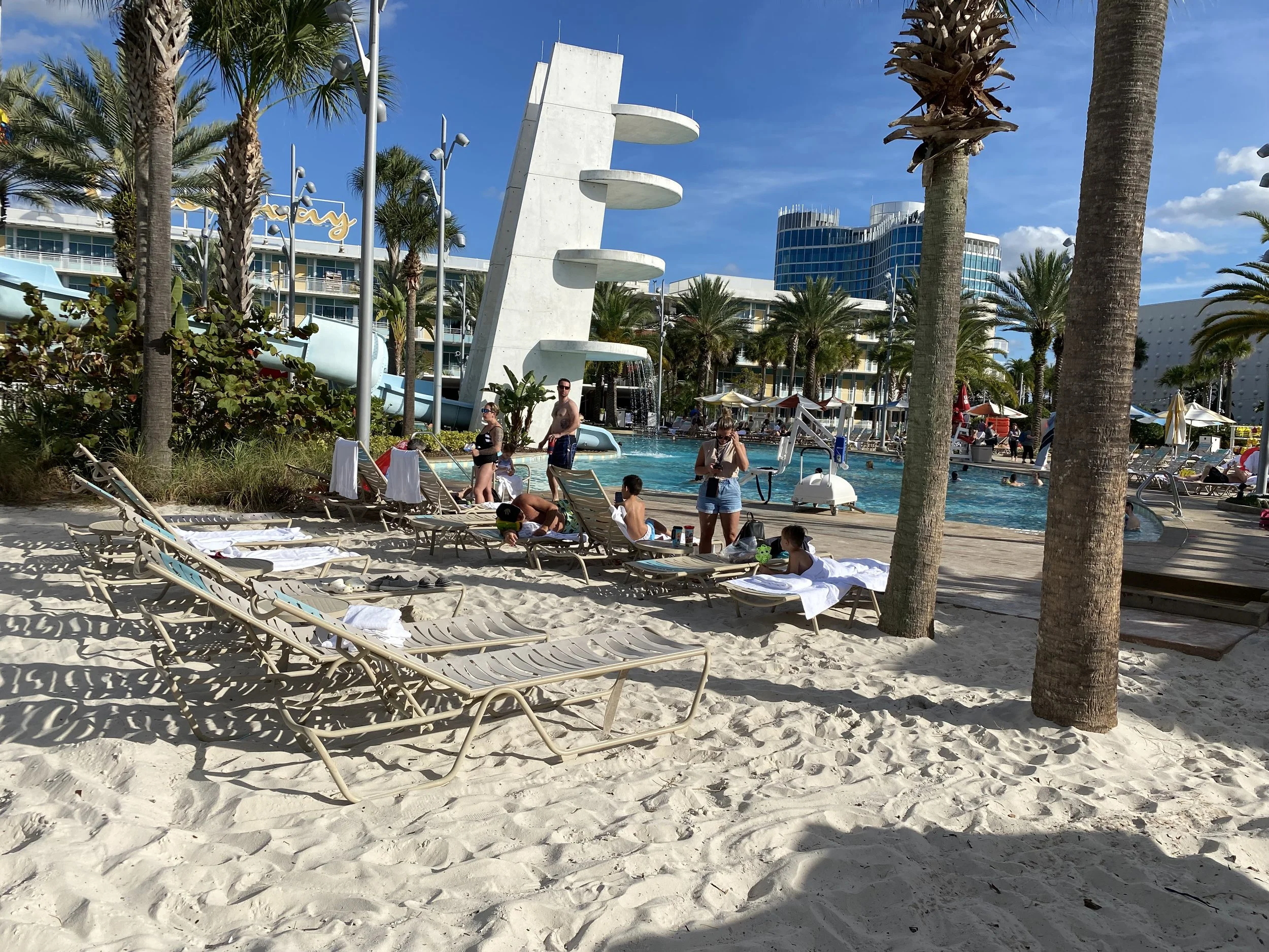 Pool area at a sandy beach with lounge chairs, palm trees, and a water slide in the background. People are relaxing and swimming.