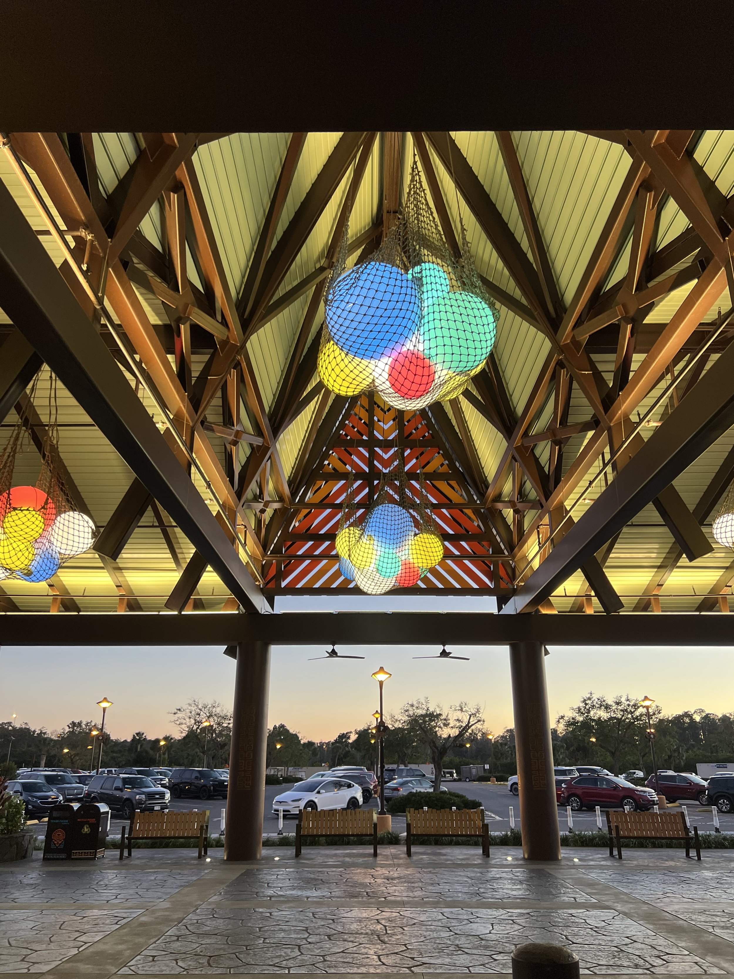 Colorful spherical lights hanging from a vaulted ceiling in an outdoor space, with a parking lot and trees visible in the background.
