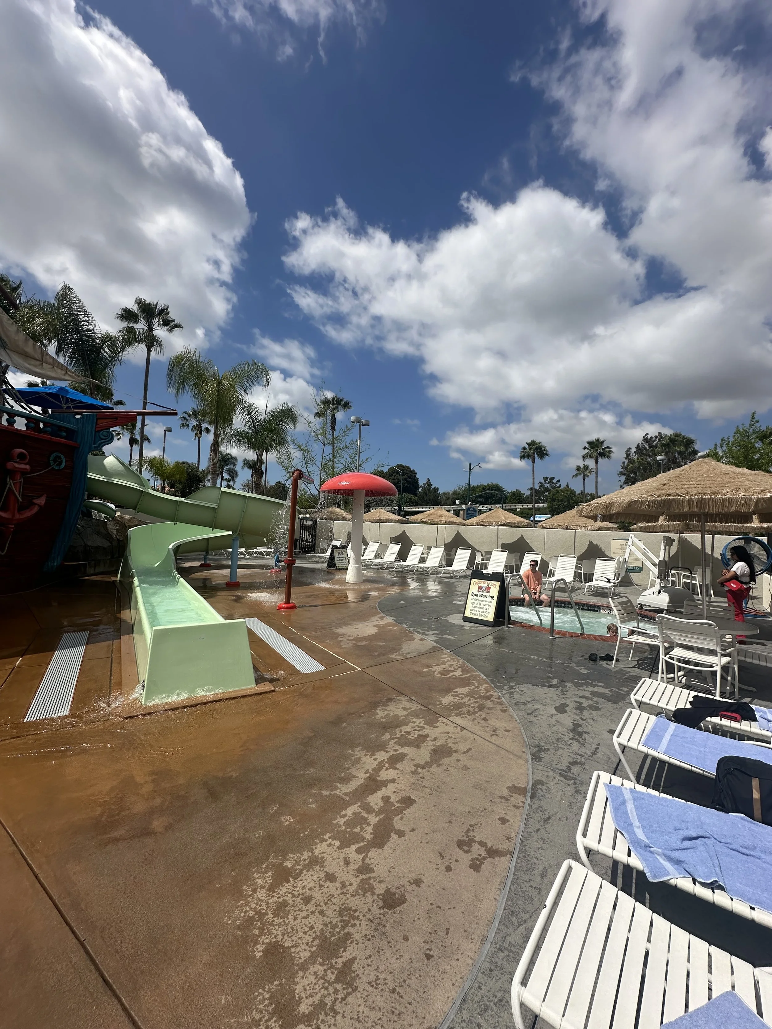 Water park with a waterslide, pool, lounge chairs, umbrellas, and a clear sky with scattered clouds.