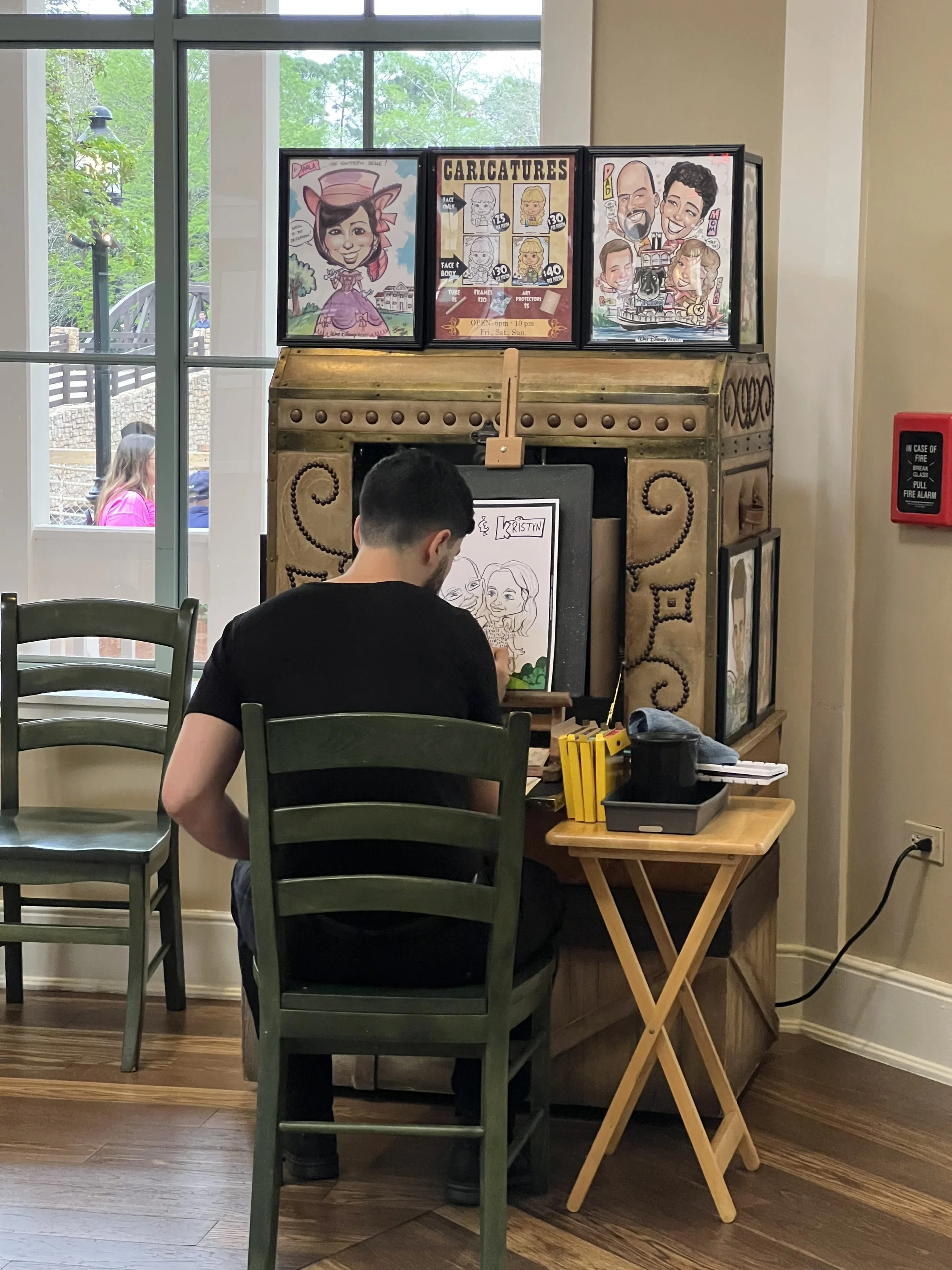A young man sitting at a small wooden table drawing a caricature of two women at a restaurant or cafe. Behind him is a large screen with caricature art samples and a caricature booth.