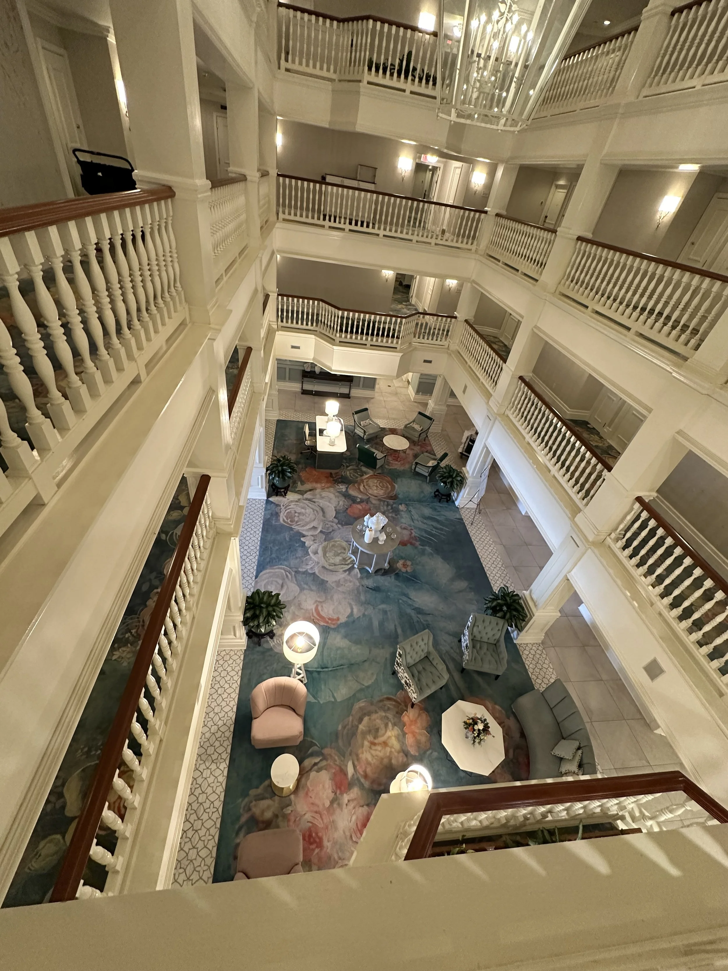 View of a hotel lobby from an upper floor, showing multiple levels with white railings, a colorful floral carpet, and various seating areas with chairs, tables, and decorative lamps.