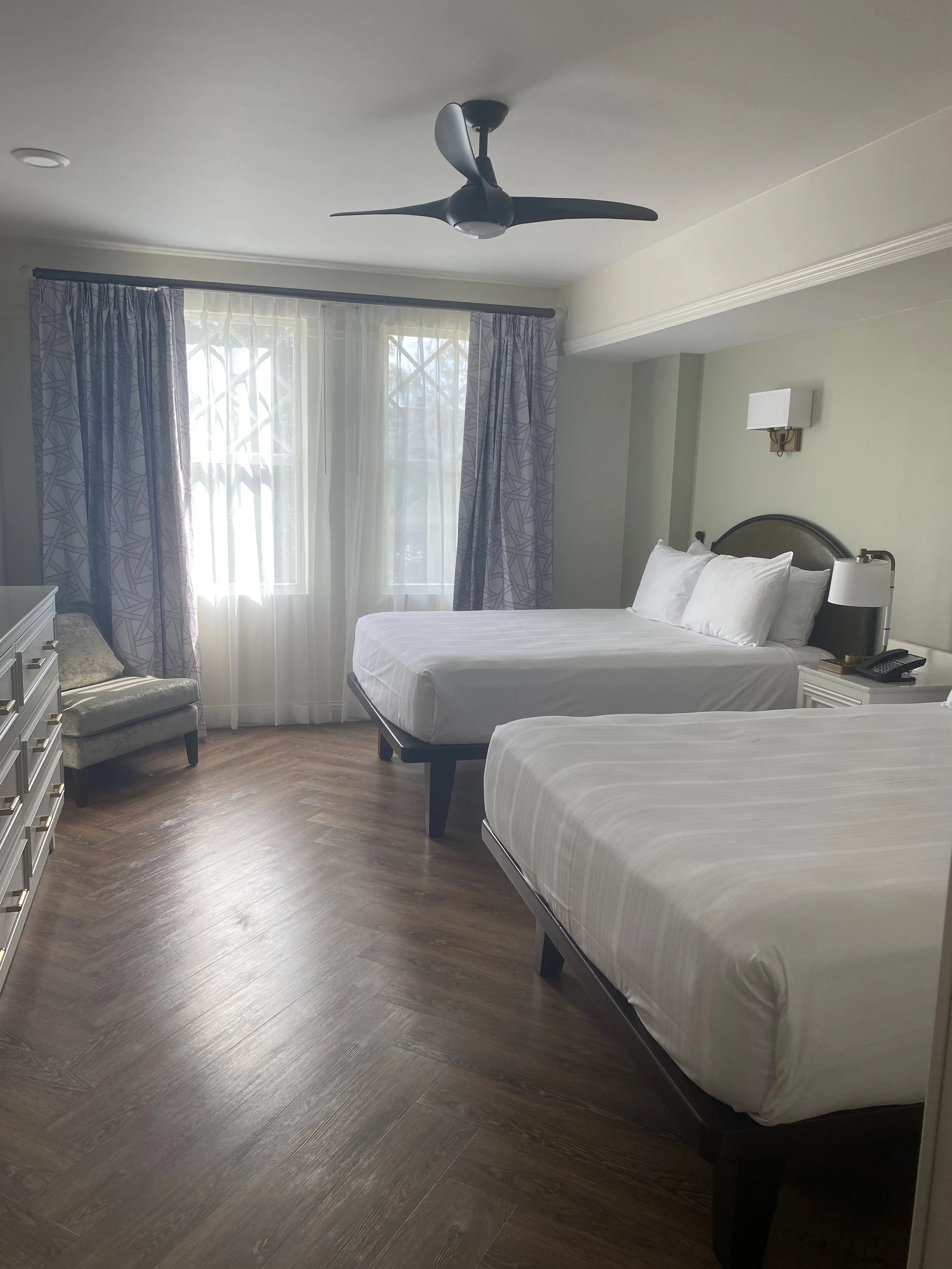A hotel room with two beds, a window with sheer curtains and patterned drapes, a ceiling fan, a dresser, a small chair, and a wall-mounted lamp.