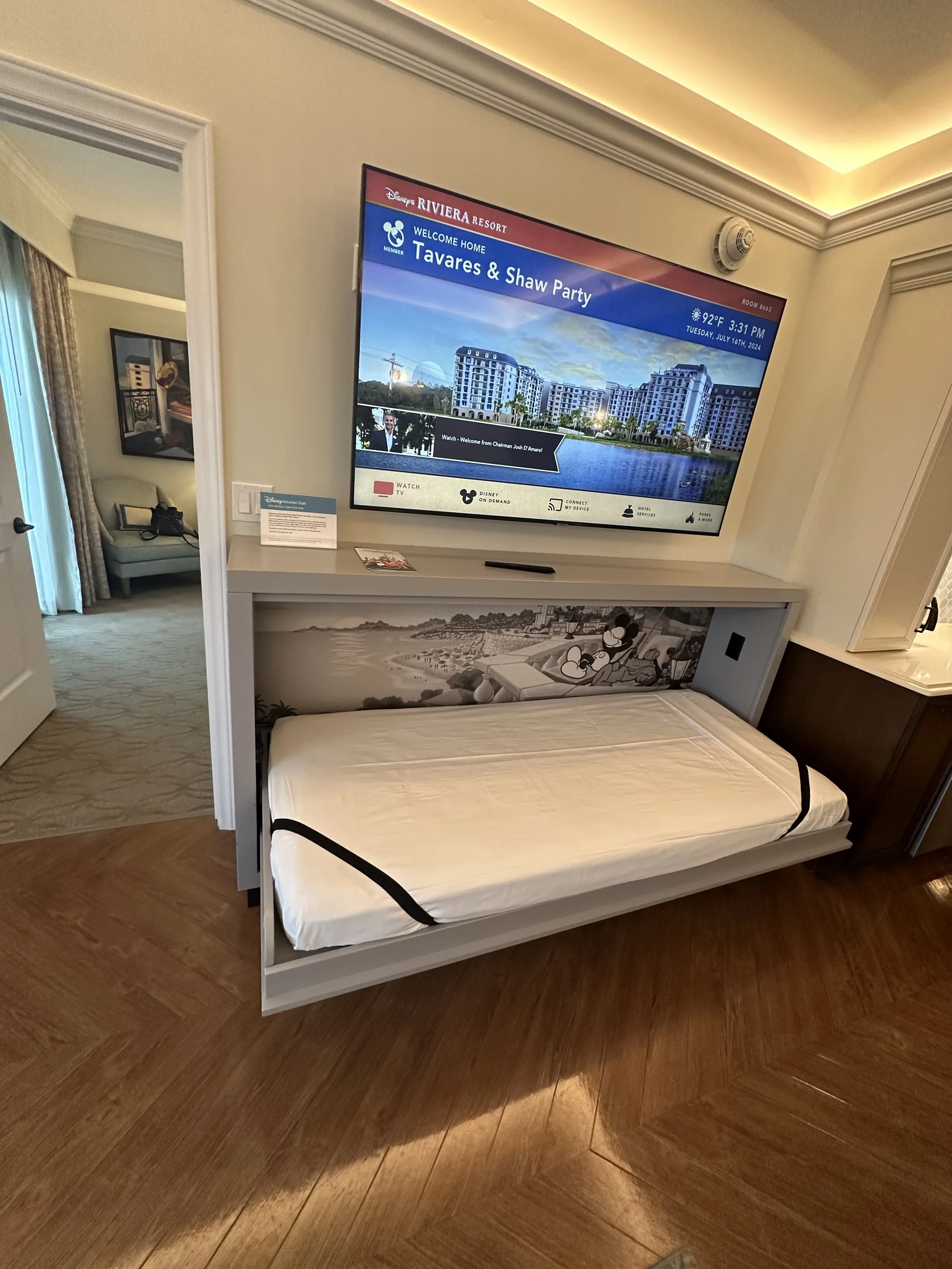 Pull-out sofa bed underneath a wall-mounted TV in a hotel room, with a nightstand and a partial view of another room.