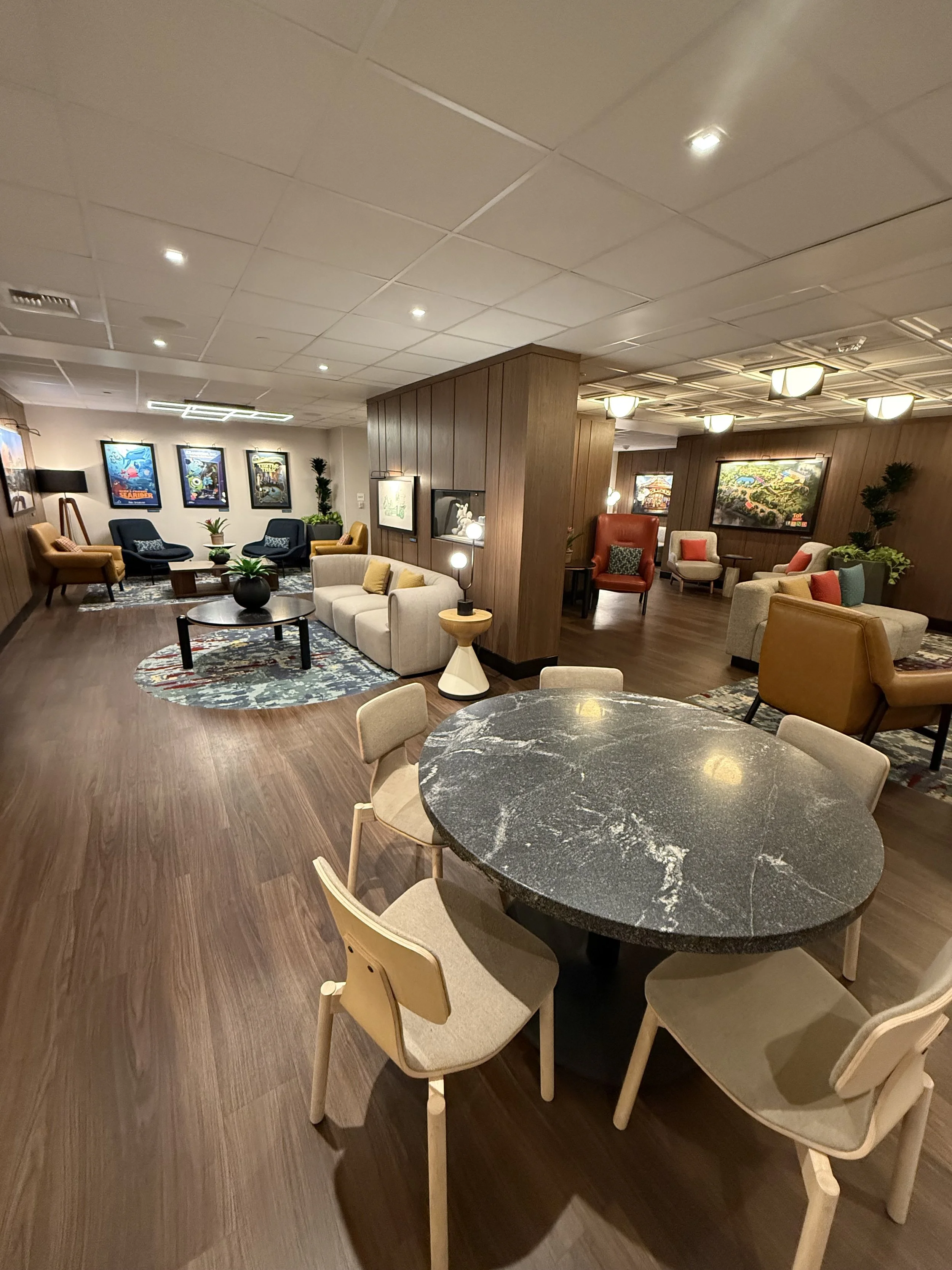 A spacious lounge area with wooden flooring and a variety of colorful armchairs and sofas arranged around coffee tables. The walls are decorated with framed artwork, and there are potted plants for added greenery. Overhead lighting illuminates the ro