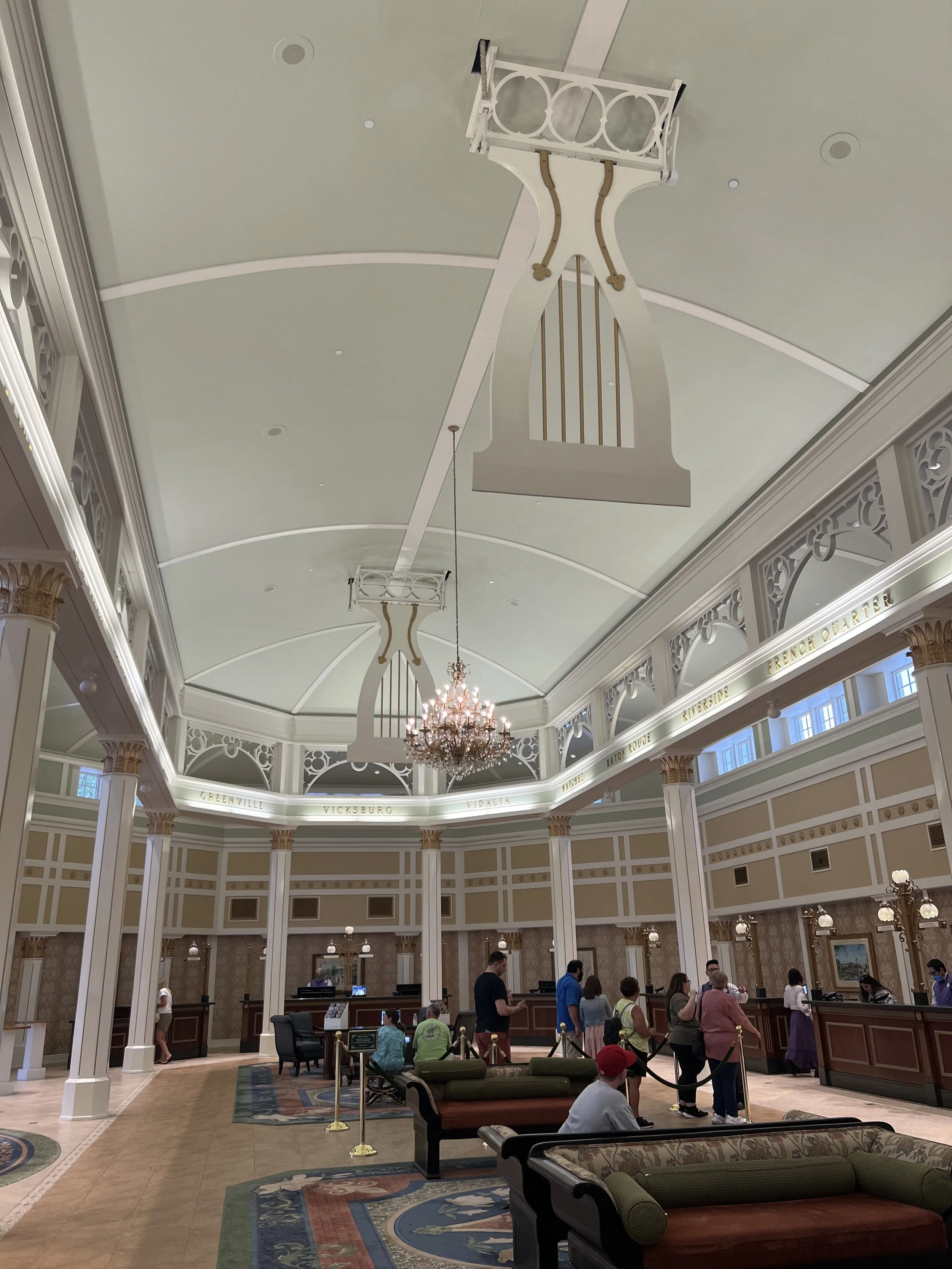 A hotel or resort lobby with a high ceiling, ornate chandelier, and decorative railings. People are standing in line at a reception desk, and seating areas with sofas are in the foreground.