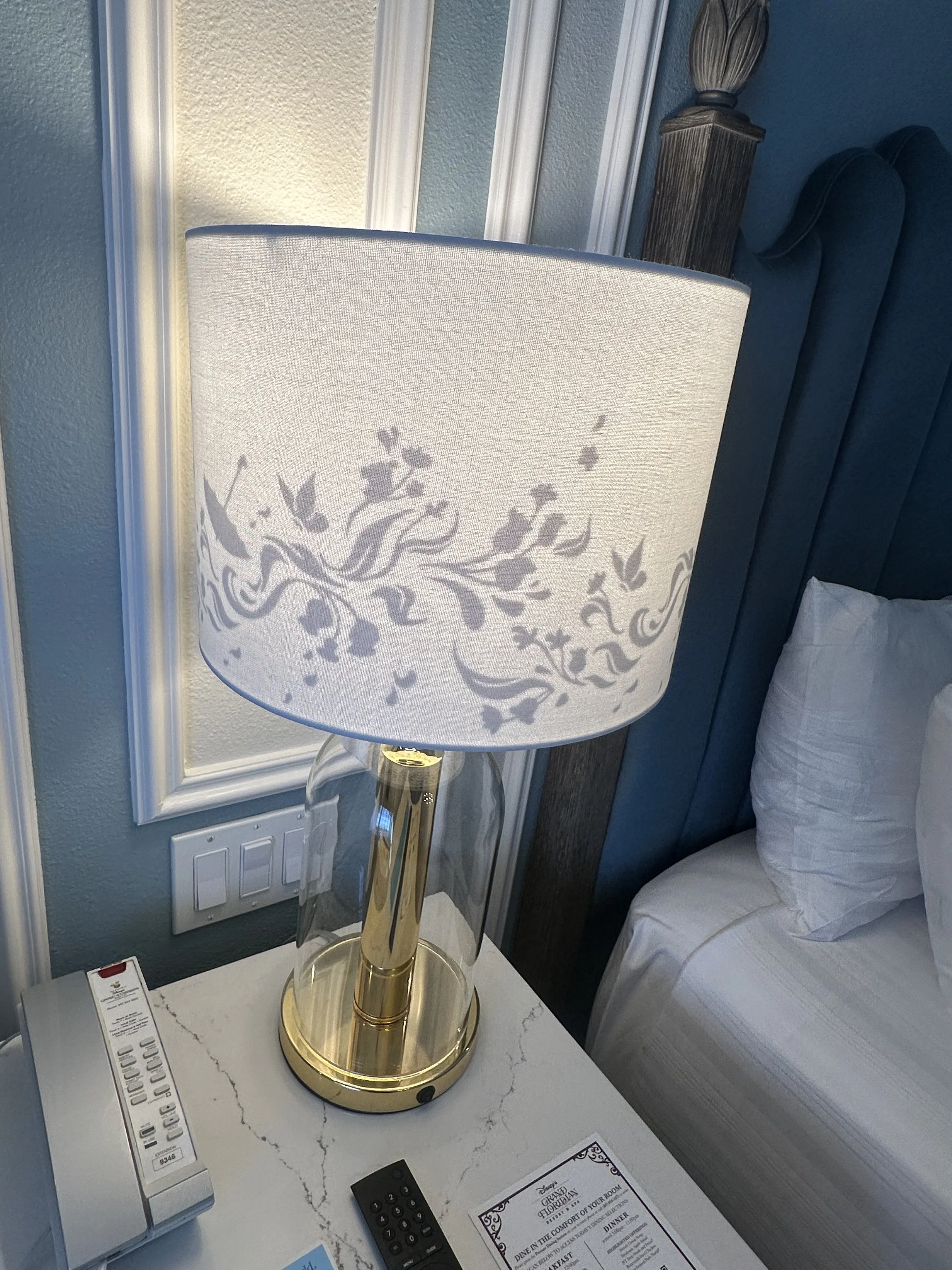 A bedside lamp with a white lampshade featuring a floral pattern, on a white marble nightstand next to a bed with white linens and pillows, a remote control, and a hotel directory, in a hotel room with light green walls and white trim.