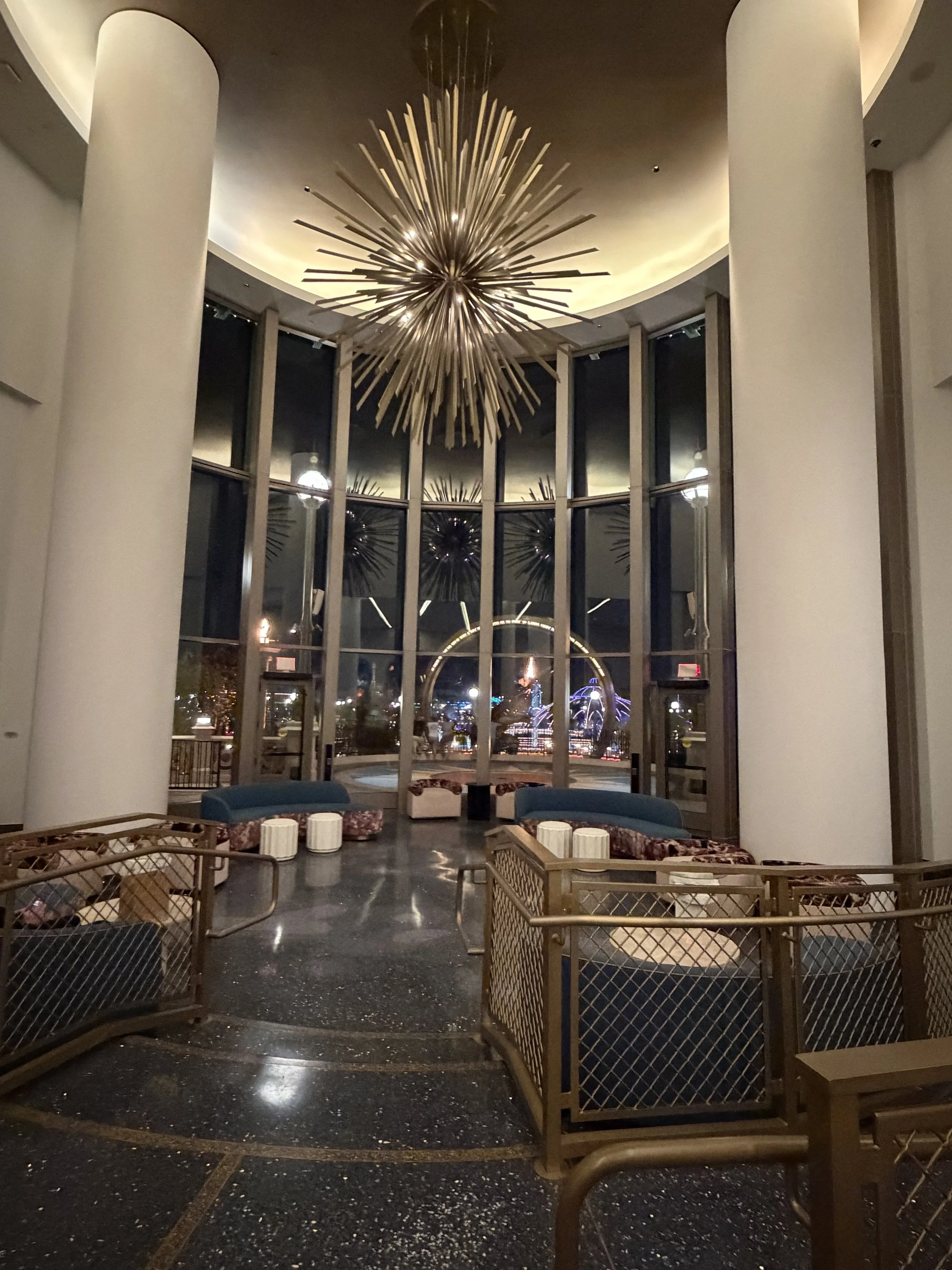 Interior of a modern building lobby with large decorative chandelier, tall windows showcasing a Ferris wheel outside at night, and seating arrangements.