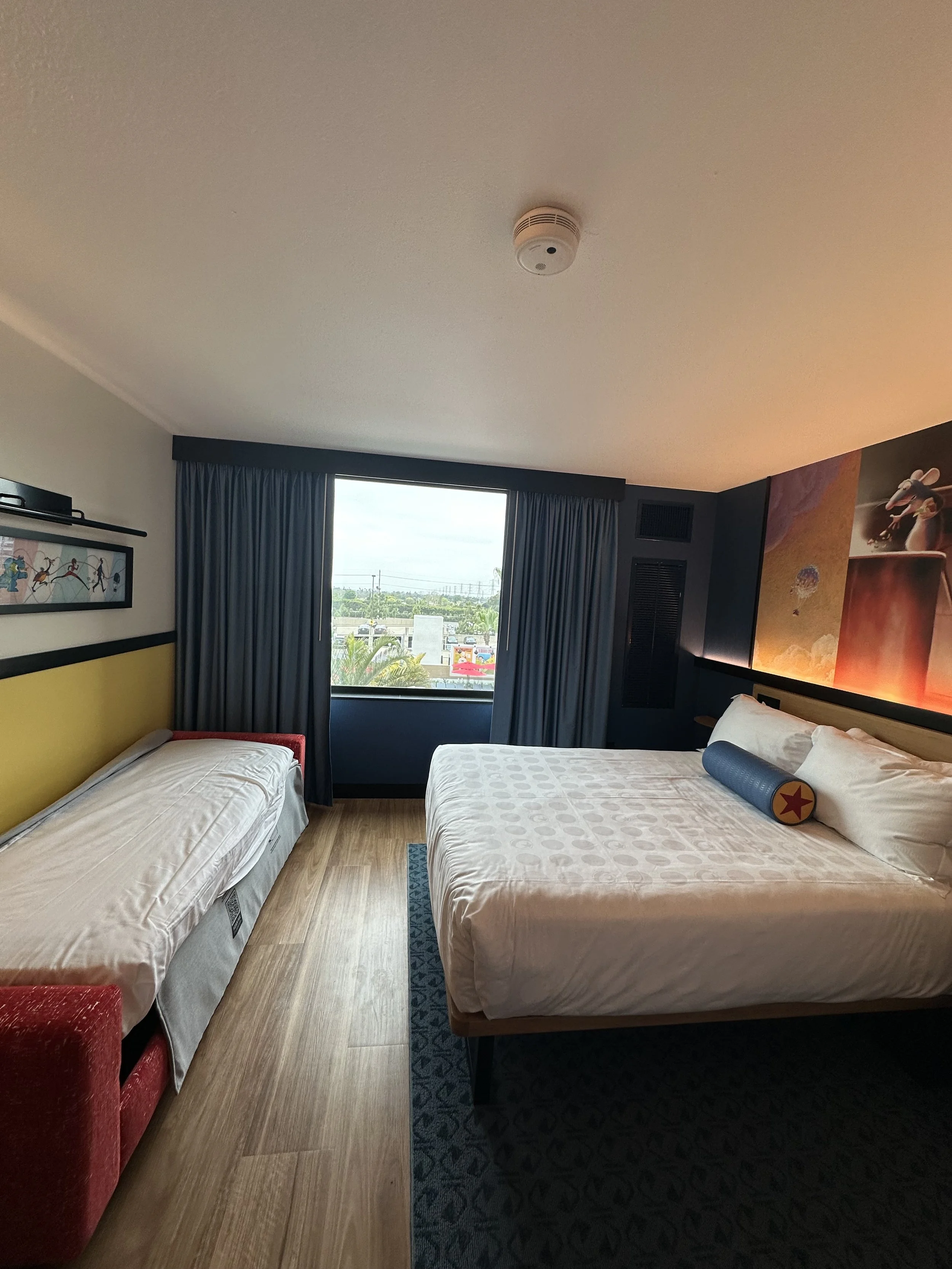 A hotel room with a large bed and a smaller bed, both with white bedding, near a window with blue curtains. The room has wooden floors and colorful wall art above the beds.