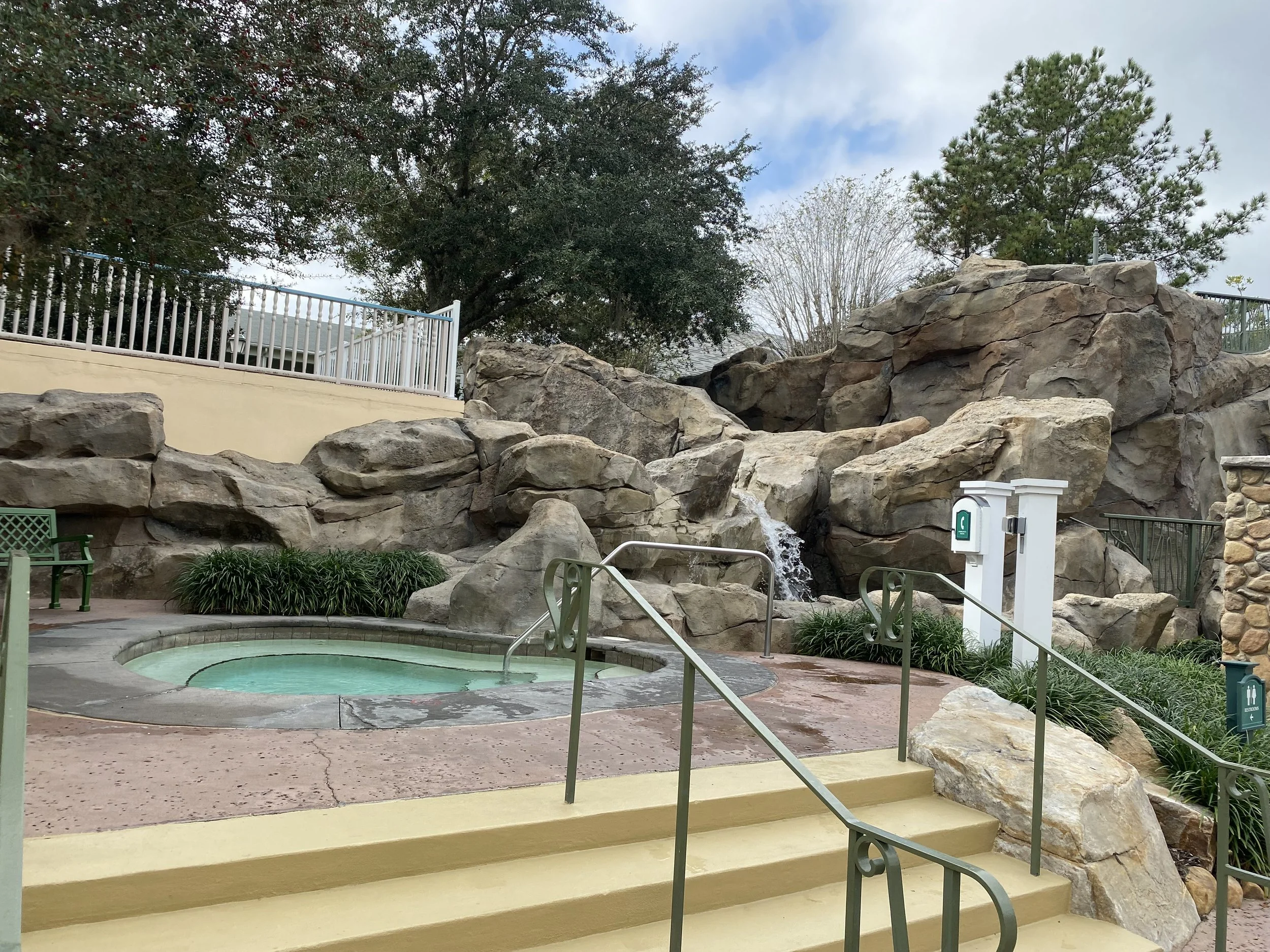 Outdoor hot tub area with a small waterfall flowing over rocks, surrounded by greenery and rocks, with stairs and handrails leading down to the area.