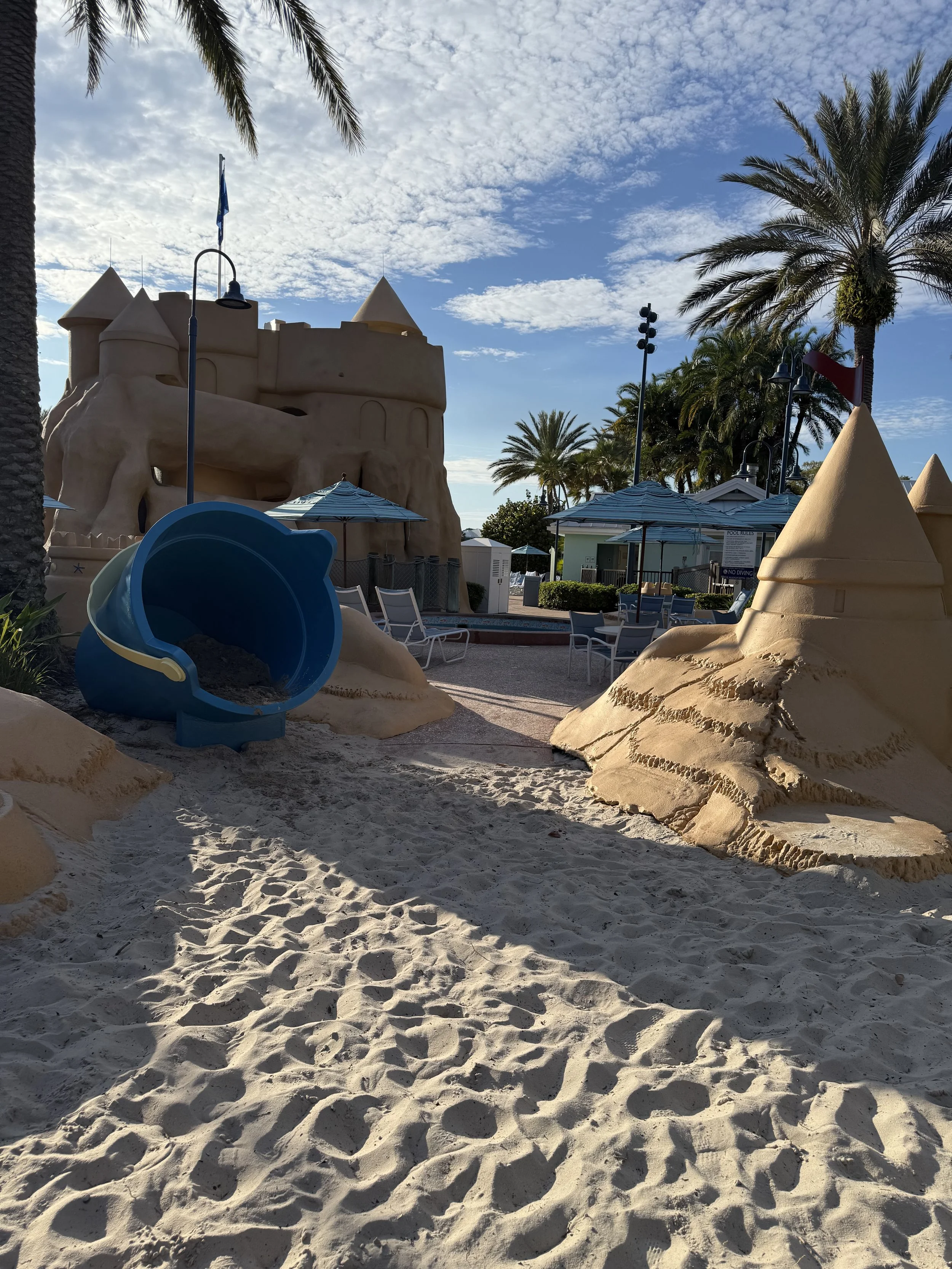 Empty children’s playground with sand, a blue sandbox shaped like a bucket, sandcastles, and shade umbrellas, surrounded by palm trees under a partly cloudy sky.