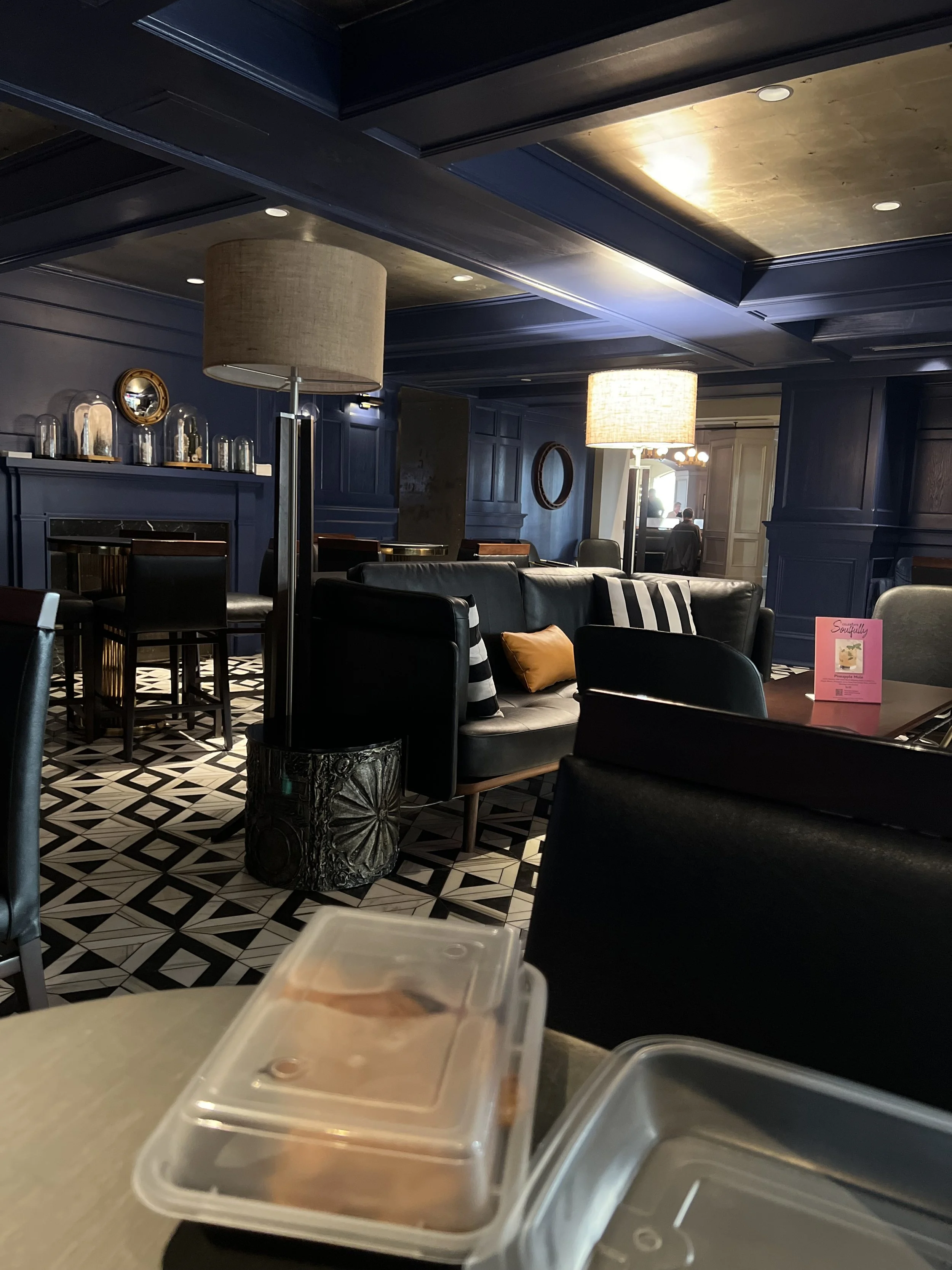 Interior of a stylish restaurant or cafe with black leather sofas, patterned black-and-white tiled floor, and contemporary lighting. There are decorative elements like mirrors and glass jars on a shelf and a pink menu card on the table.