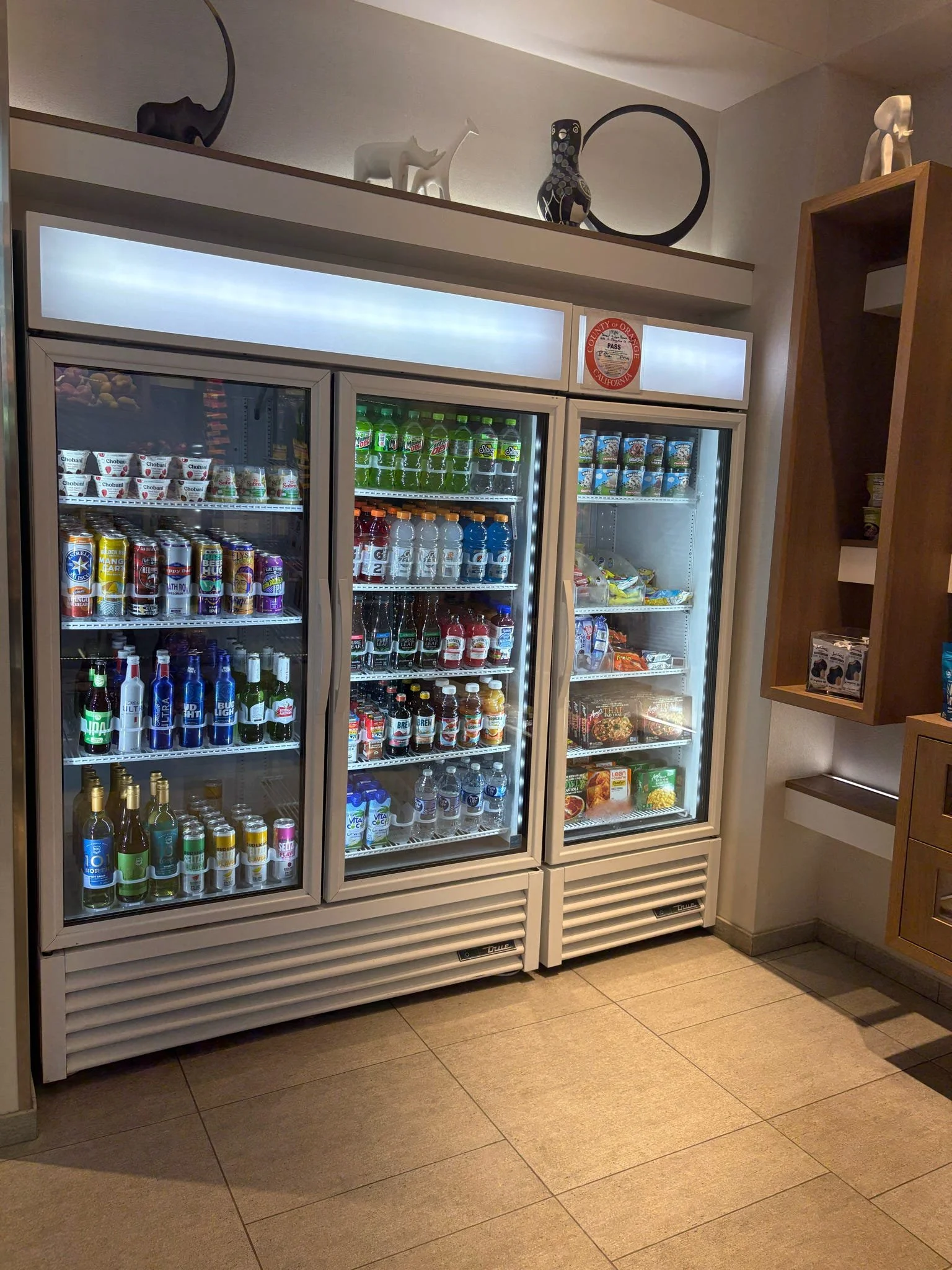 Three glass-door refrigerators filled with beverages, including soda, water, and energy drinks, in a convenience store. Decor with elephant and animal sculptures on a shelf above the refrigerators.