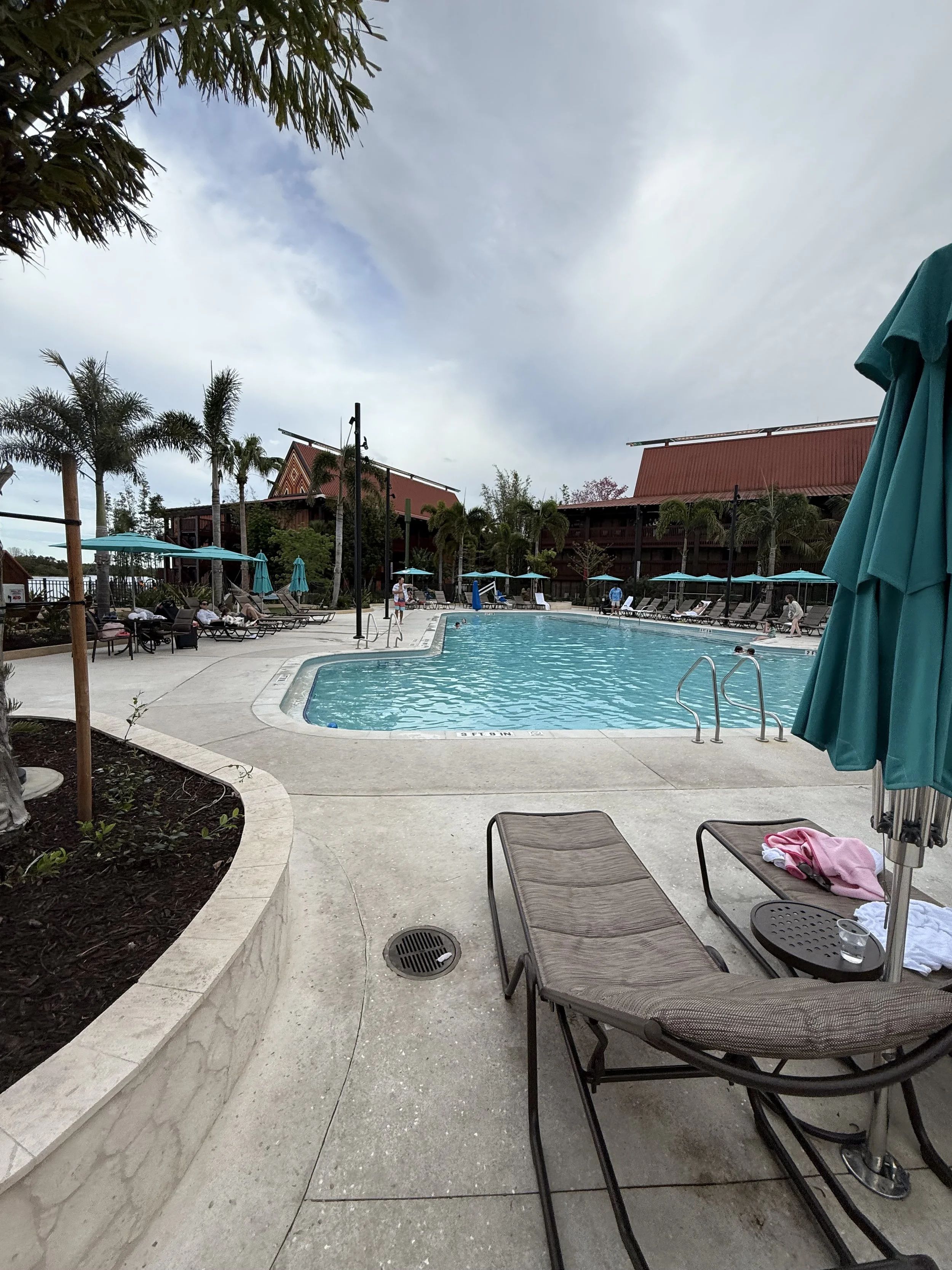 An outdoor swimming pool with lounge chairs, umbrellas, and palm trees surrounding it, under a partly cloudy sky.