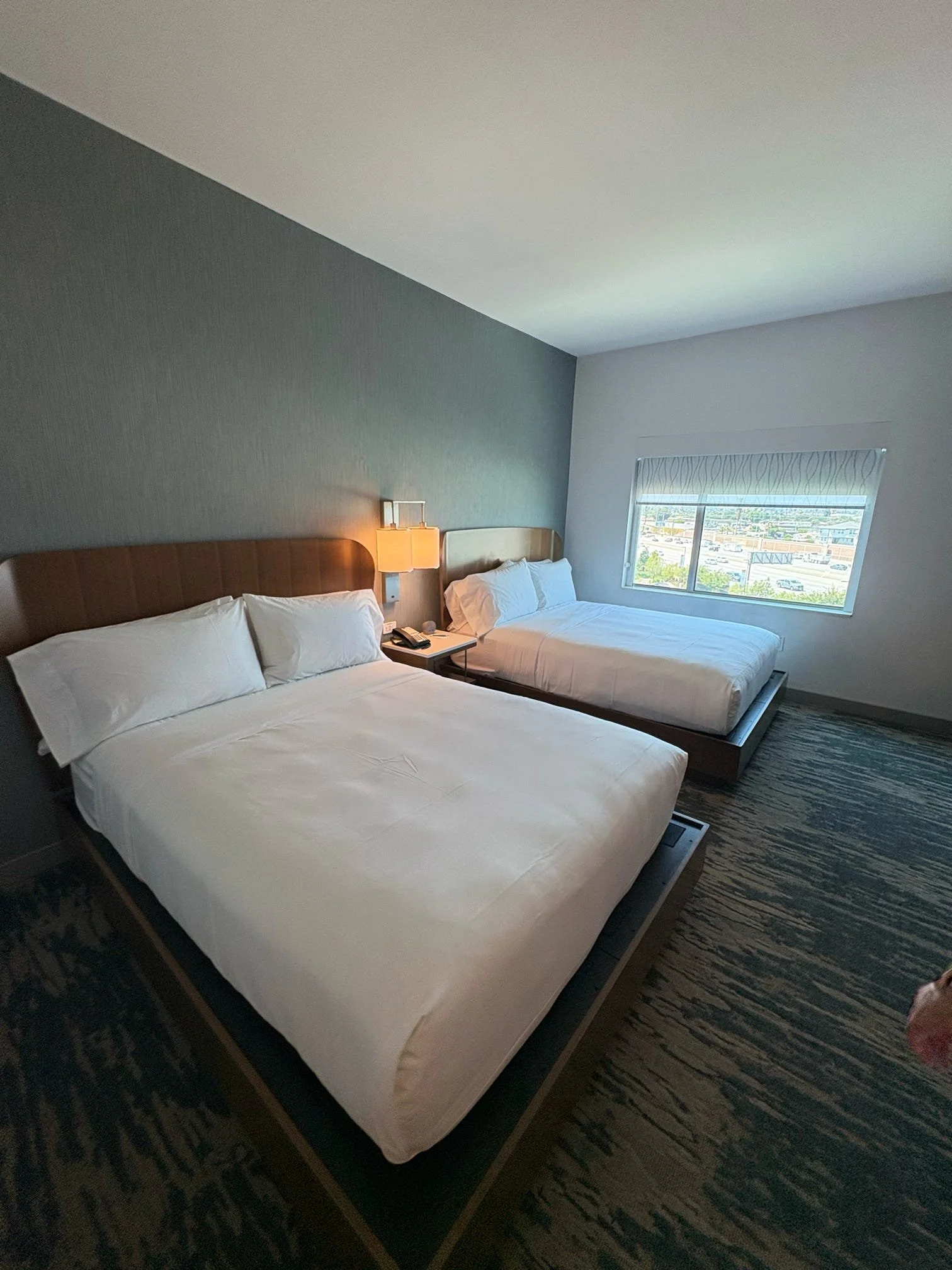 A hotel room with two queen-sized beds, a window with a city view, and a small nightstand between the beds.