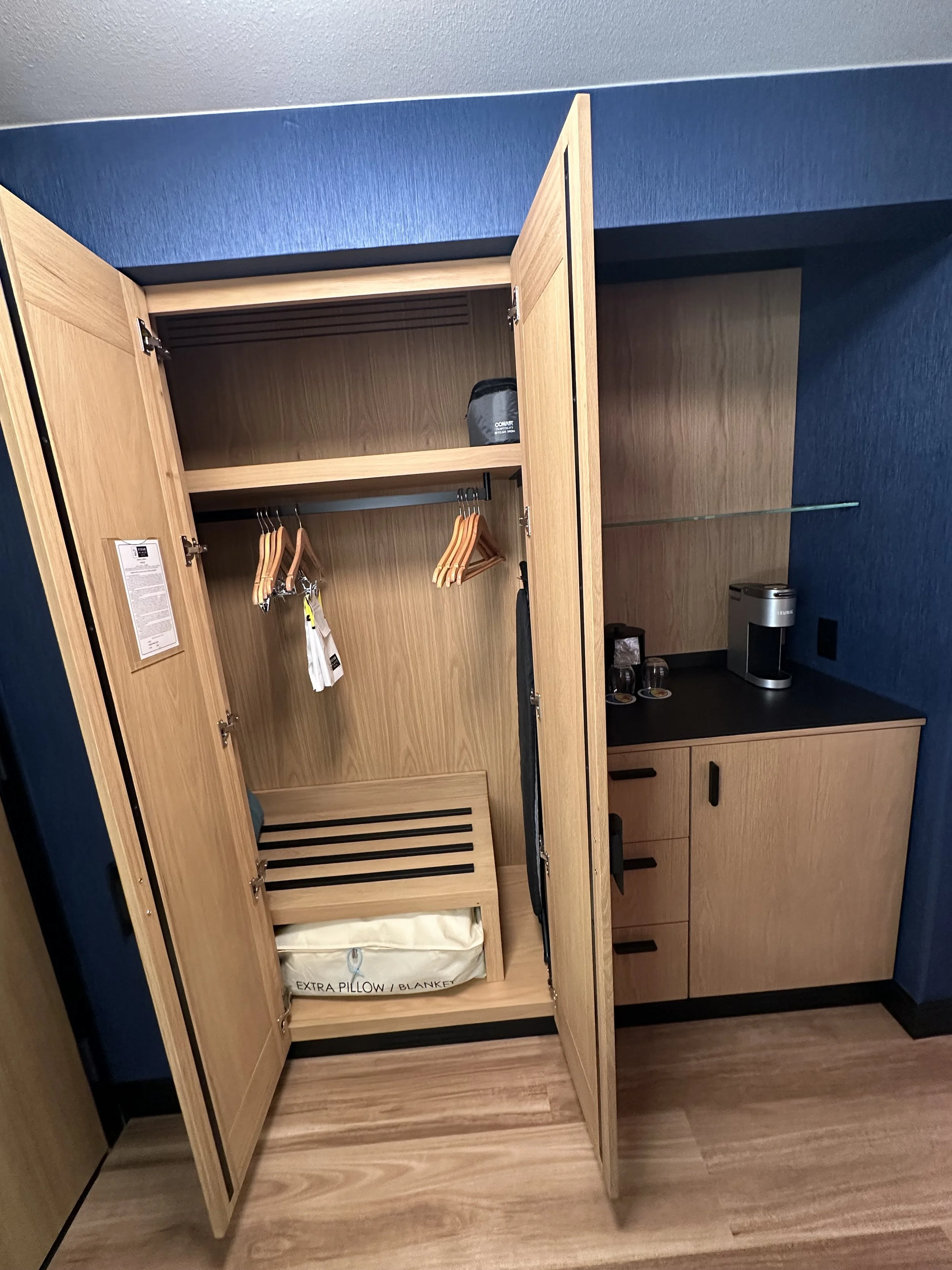 Open wooden closet with clothing hangers, a bag, a shelf, a bed, and a wrapped pillow inside, beside a black countertop with a coffee machine and cups on a blue wall.