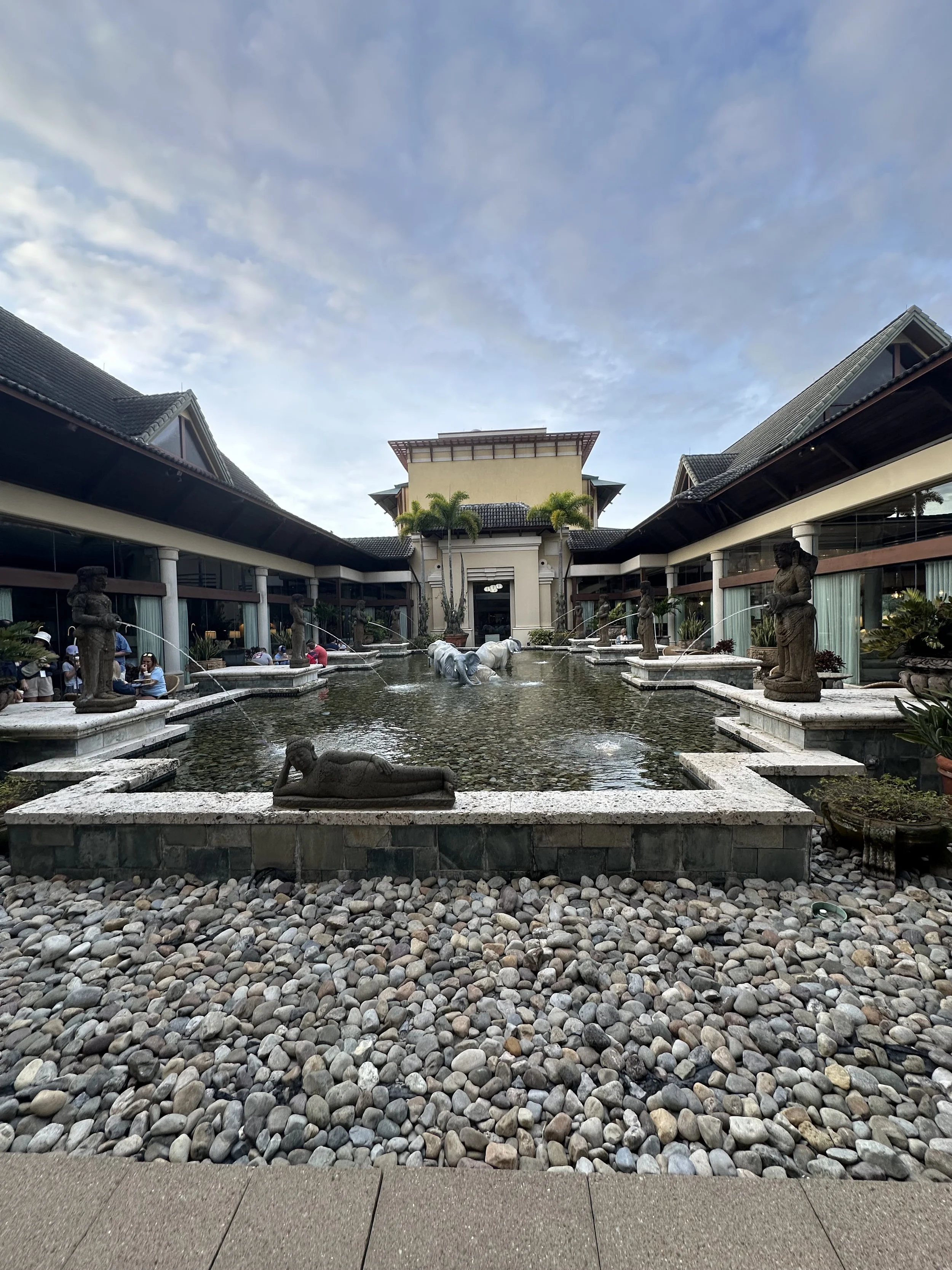 A decorative courtyard with a small pond featuring stone statues of reclining and standing figures, surrounded by pebbles, with a building and a cloudy sky in the background.