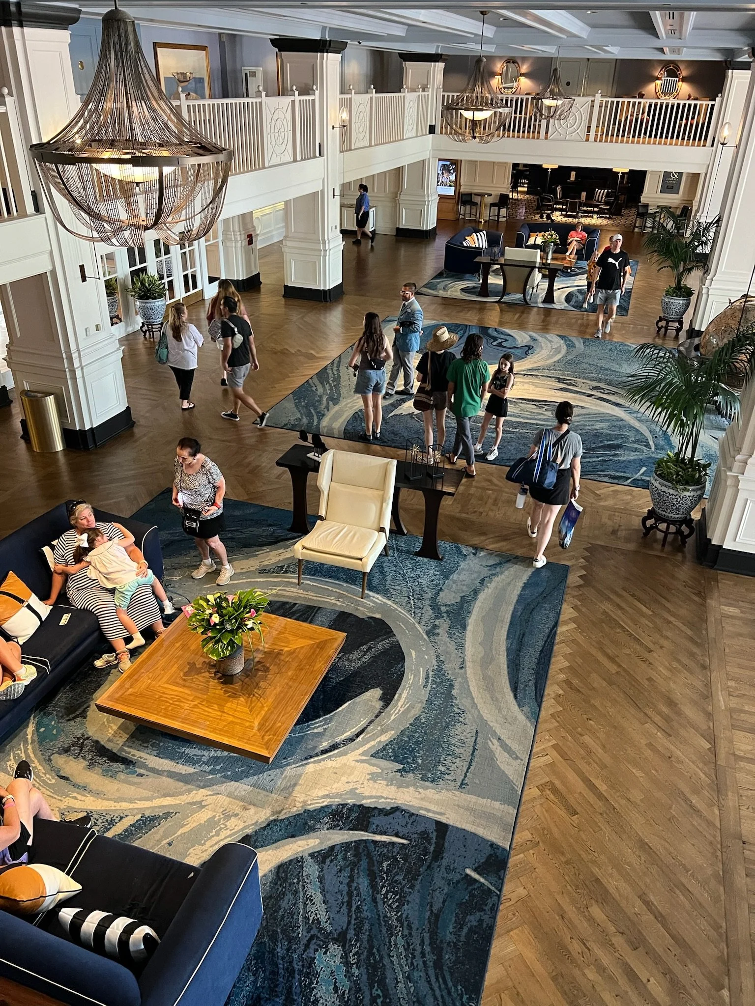 People gathered in a spacious hotel lobby with high ceilings, large chandeliers, and a patterned area rug. Some are sitting on couches, while others walk and stand, conversing or waiting. The space includes decorative potted plants, a wooden coffee t