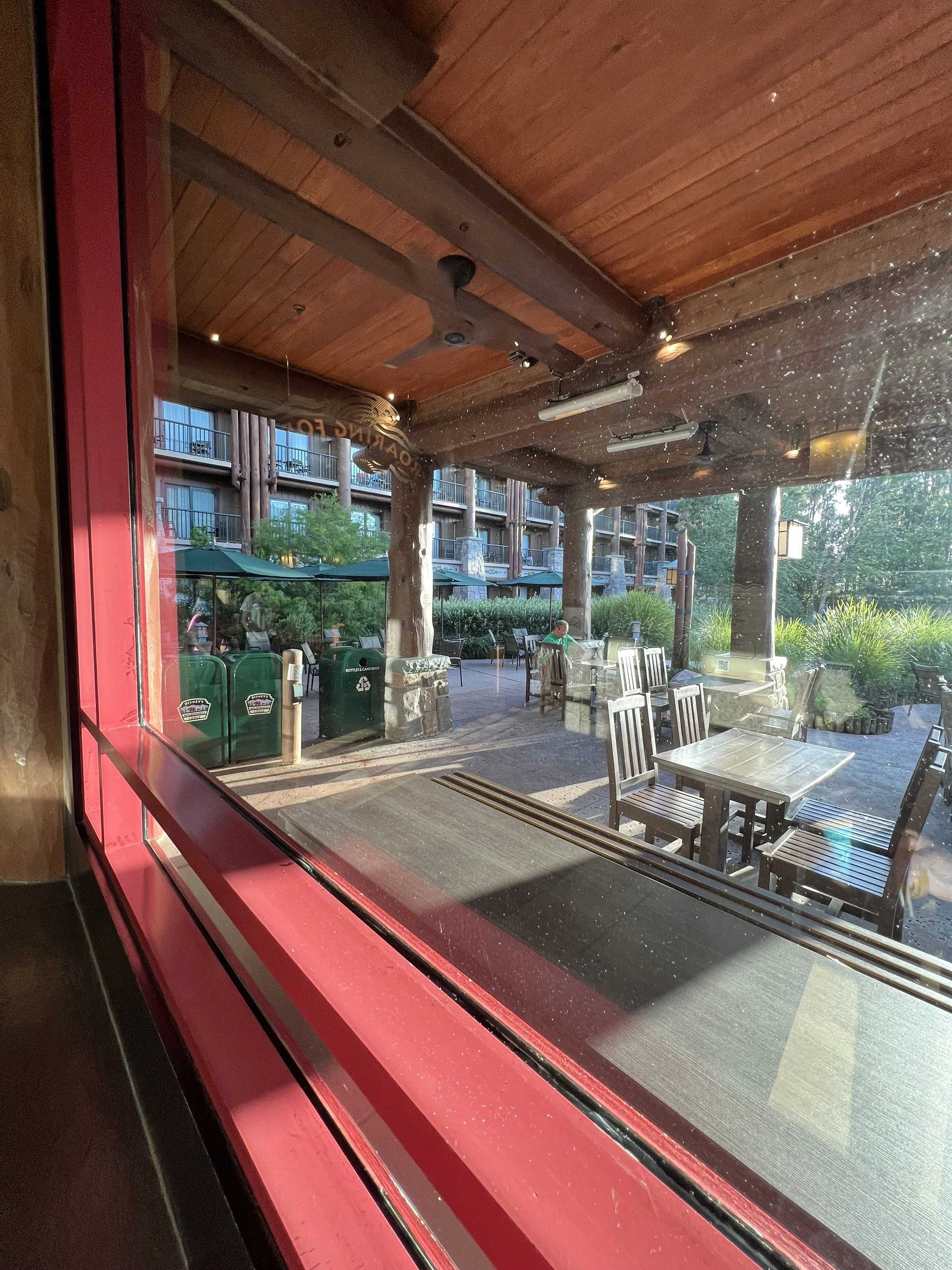 Outdoor patio seen through a window, with tables and chairs, green umbrellas, and lush greenery, at sunset.